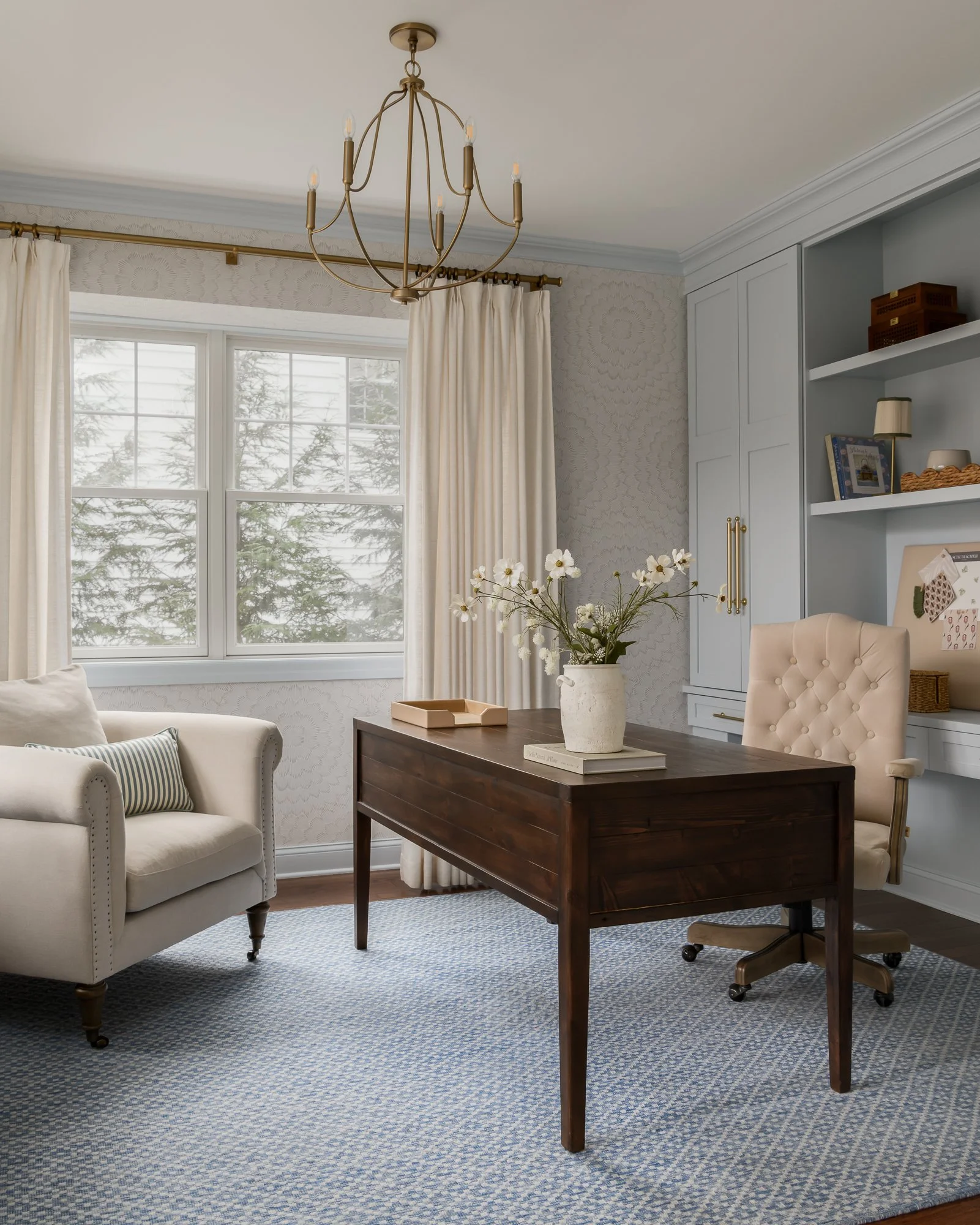 Living room with a window, beige sofa, wooden desk with a vase of white flowers, cream-colored office chair, built-in shelves, and a chandelier.