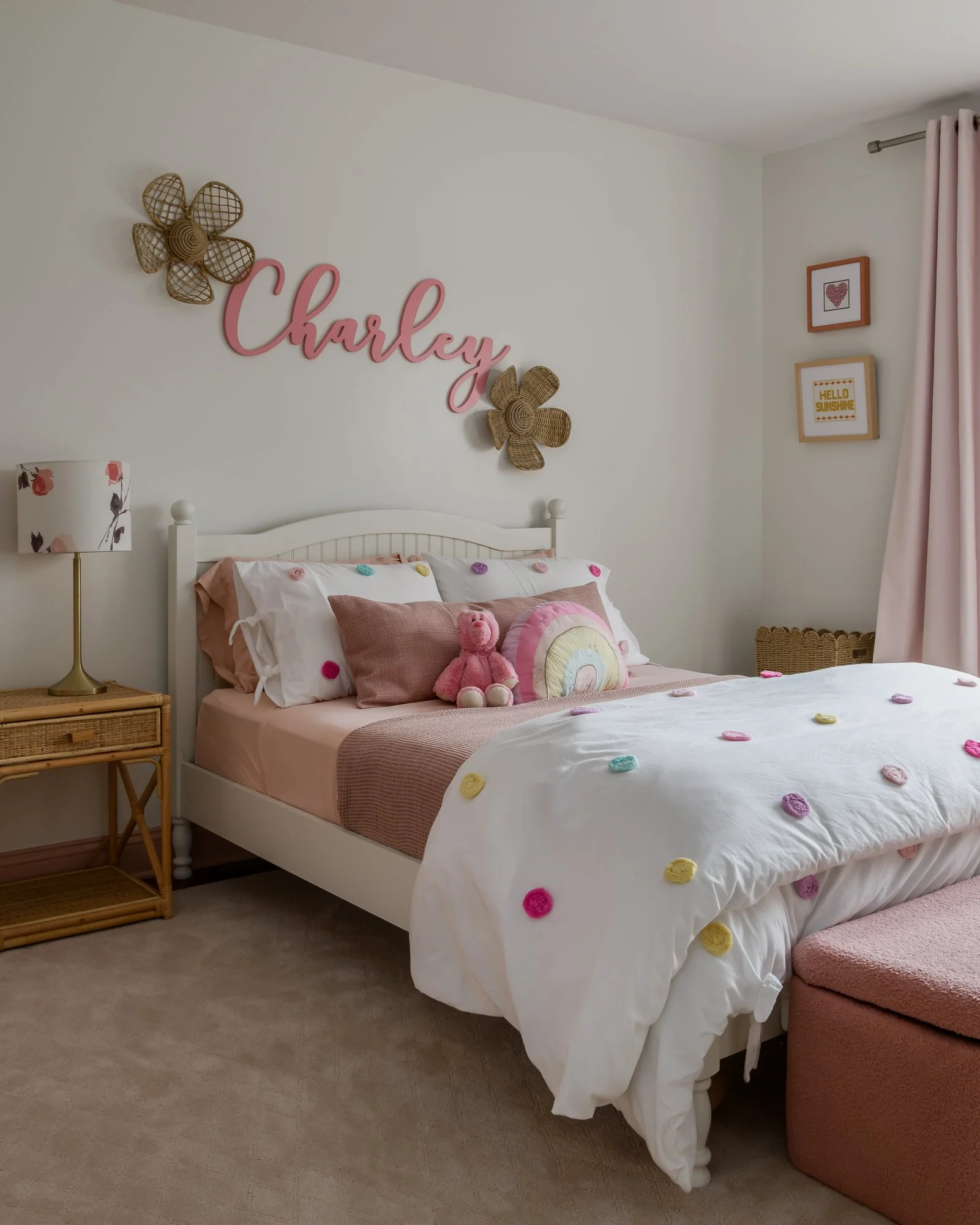 A decorated bedroom with a white bed, pink and pink accents, a ruffled pillow, a stuffed bear, and wall decorations including the name 'Charley' in pink script, woven flowers, and framed pictures.