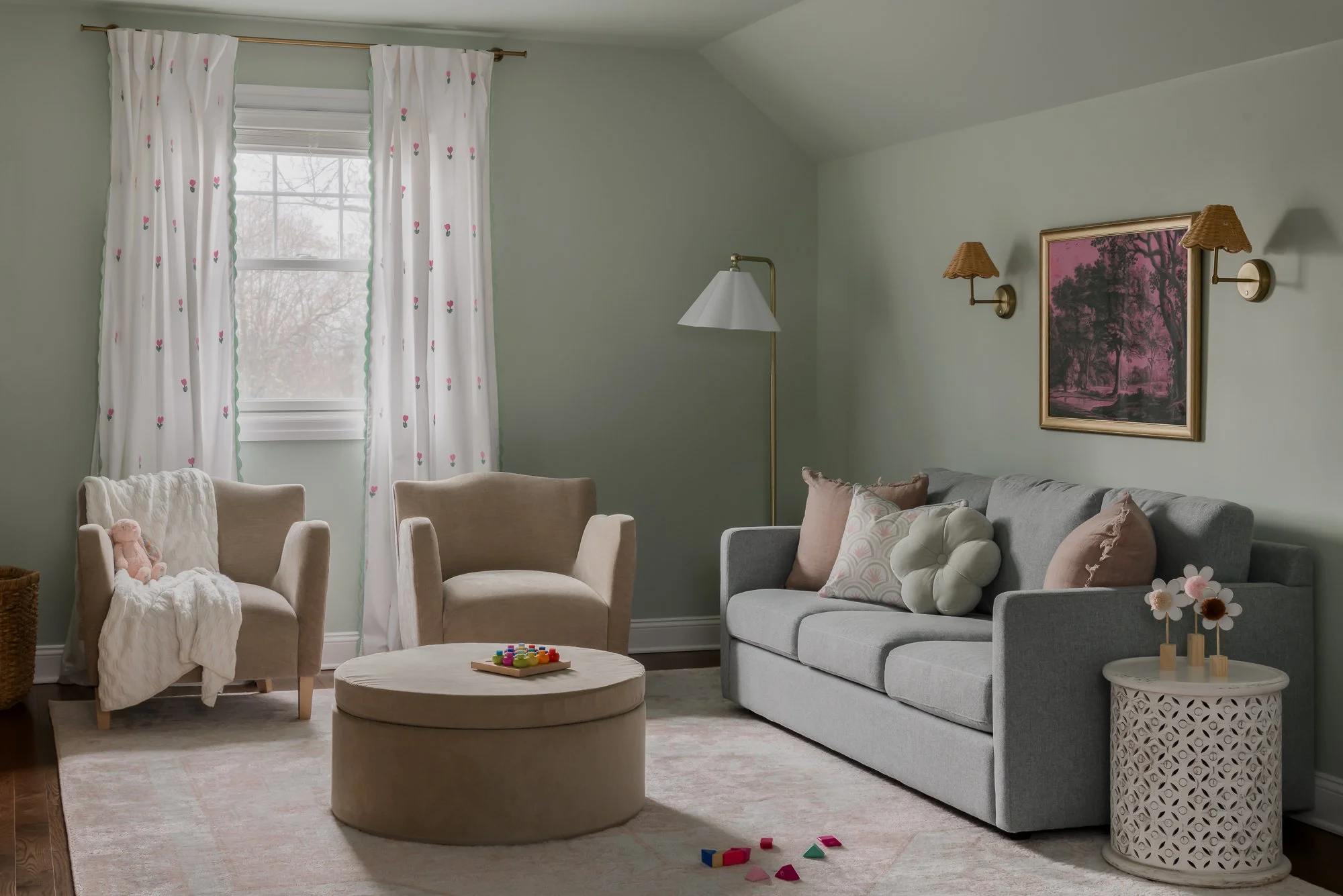 A cozy living room with beige armchairs, a gray sofa with throw pillows, a round beige ottoman, and a side table with decorative flowers. There are white curtains with small pink flowers in front of a window, with soft natural light coming in. The wa