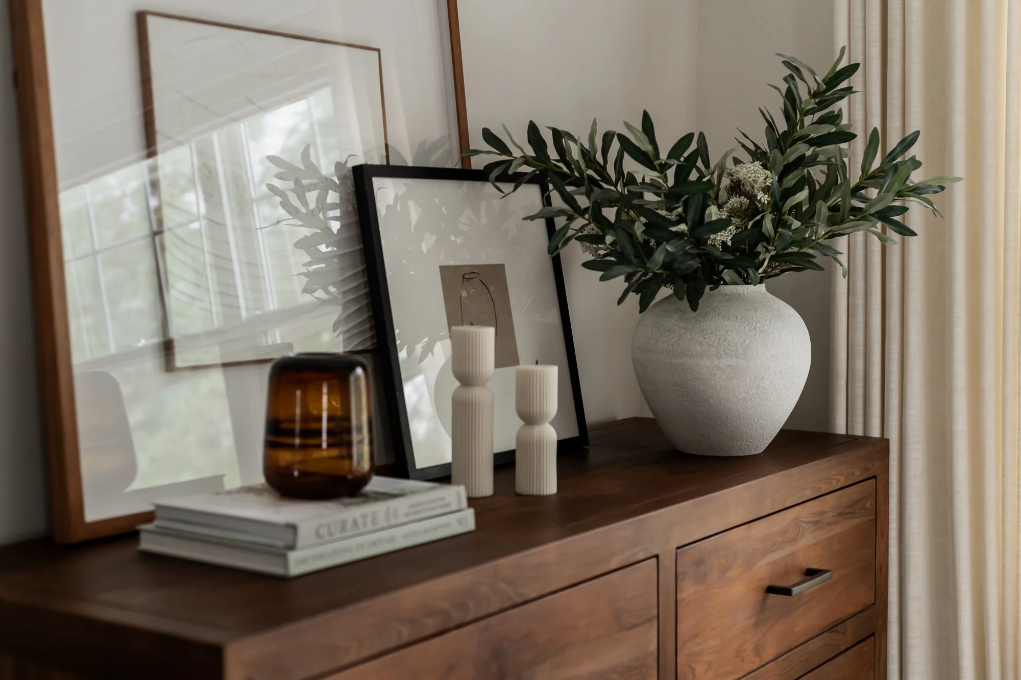 A wooden sideboard decorated with framed artwork, candles, a large vase with green foliage, and a small amber glass container.