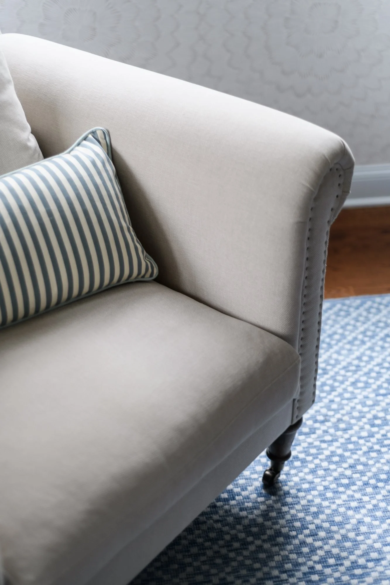 Close-up of a light-colored sofa with a blue and white striped pillow, next to a wall with textured wallpaper and a blue patterned rug on a wooden floor.