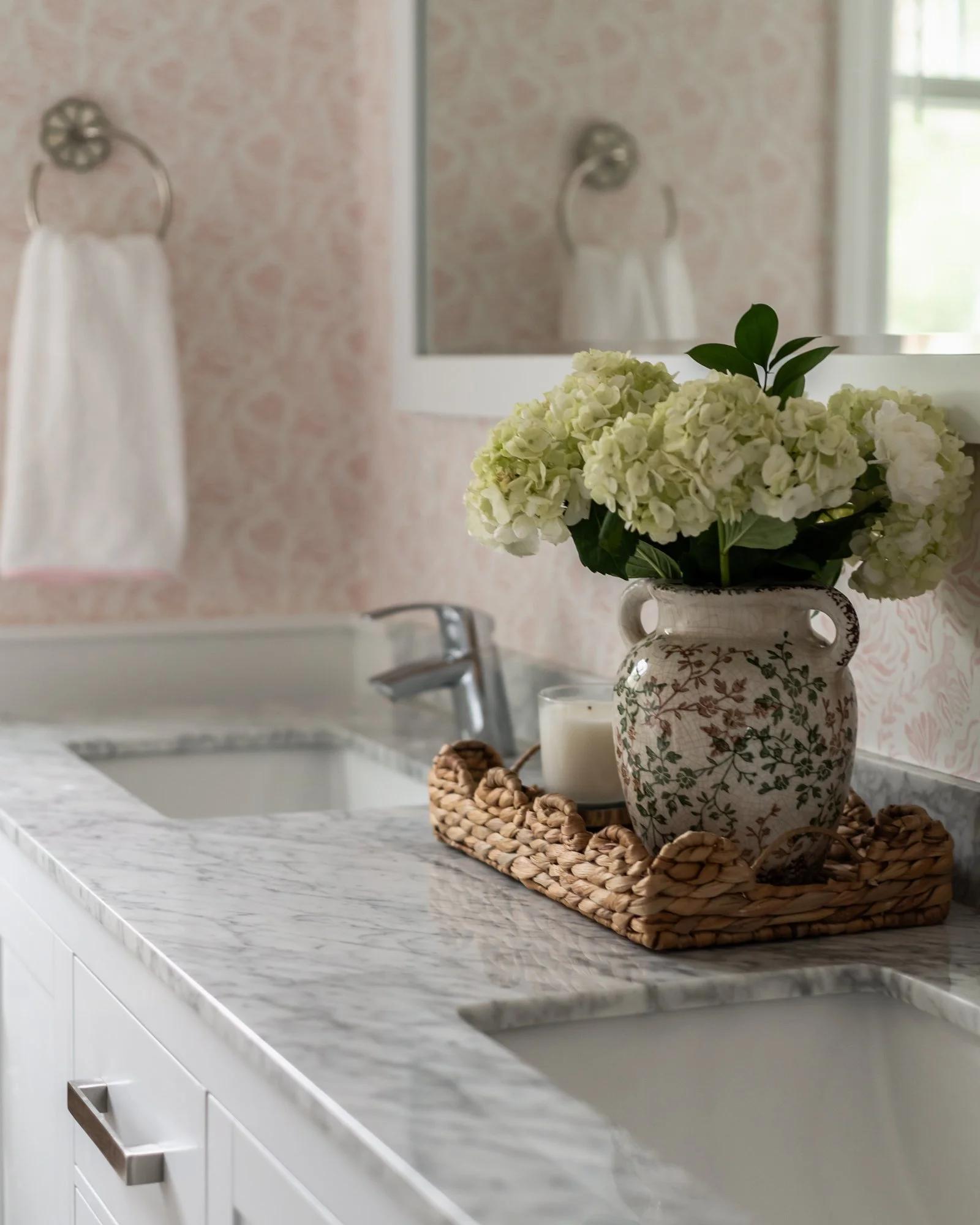 Close-up of a bathroom countertop with a decorative vase of white hydrangeas, a candle, and a woven tray, with a mirror, towel, and window in the background.