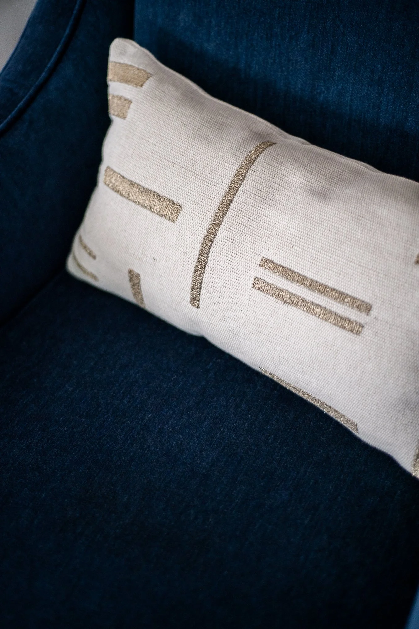 A decorative rectangular pillow with beige fabric and metallic gold lines and shapes, resting on a dark blue upholstered sofa.