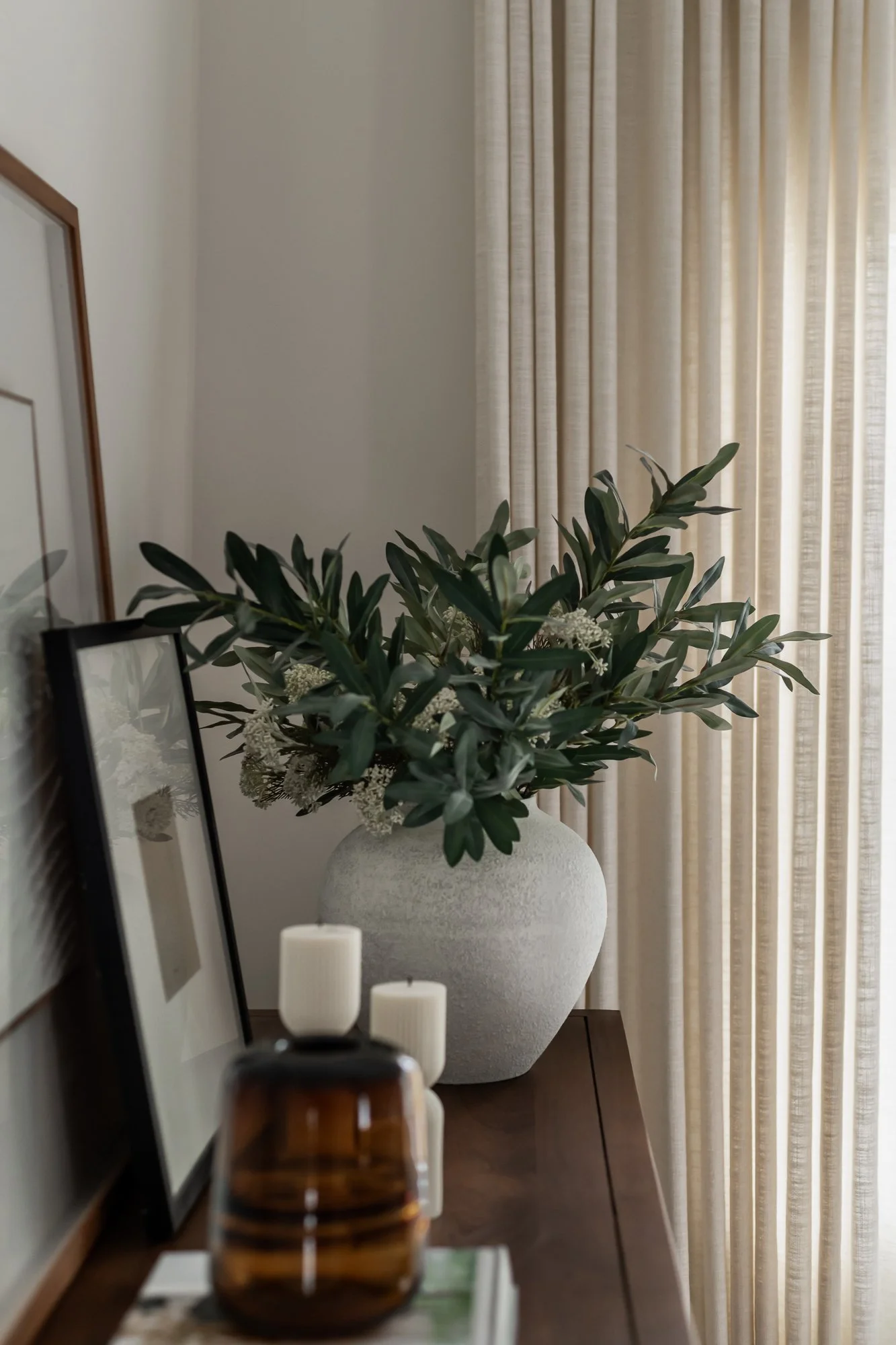 Decorative arrangement of green plants and flowers in a large white vase on a wooden table, with candles and framed artwork nearby, and cream-colored curtains in the background.