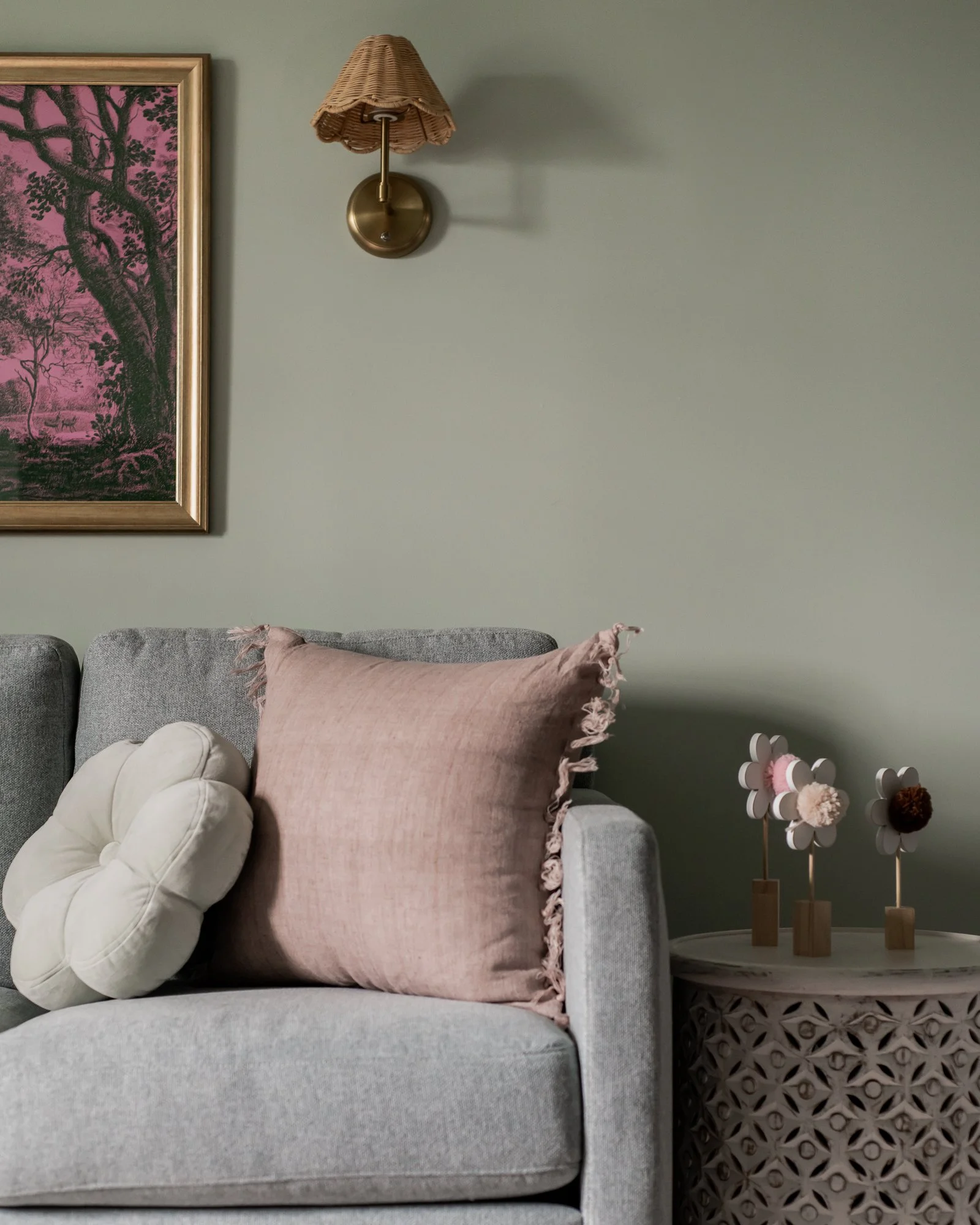 A living room scene featuring a light gray sofa with a round, cream-colored flower-shaped pillow and a pink rectangular pillow with fringed edges. To the right of the sofa, there's a white decorative table with floral-shaped ornaments on stands. The 