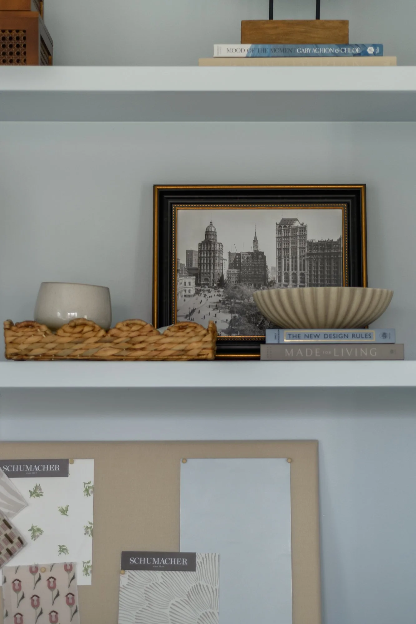 A decorative shelf with books, a framed black-and-white cityscape photograph, a beige bowl, a small beige pot, and a woven tray with desserts.
