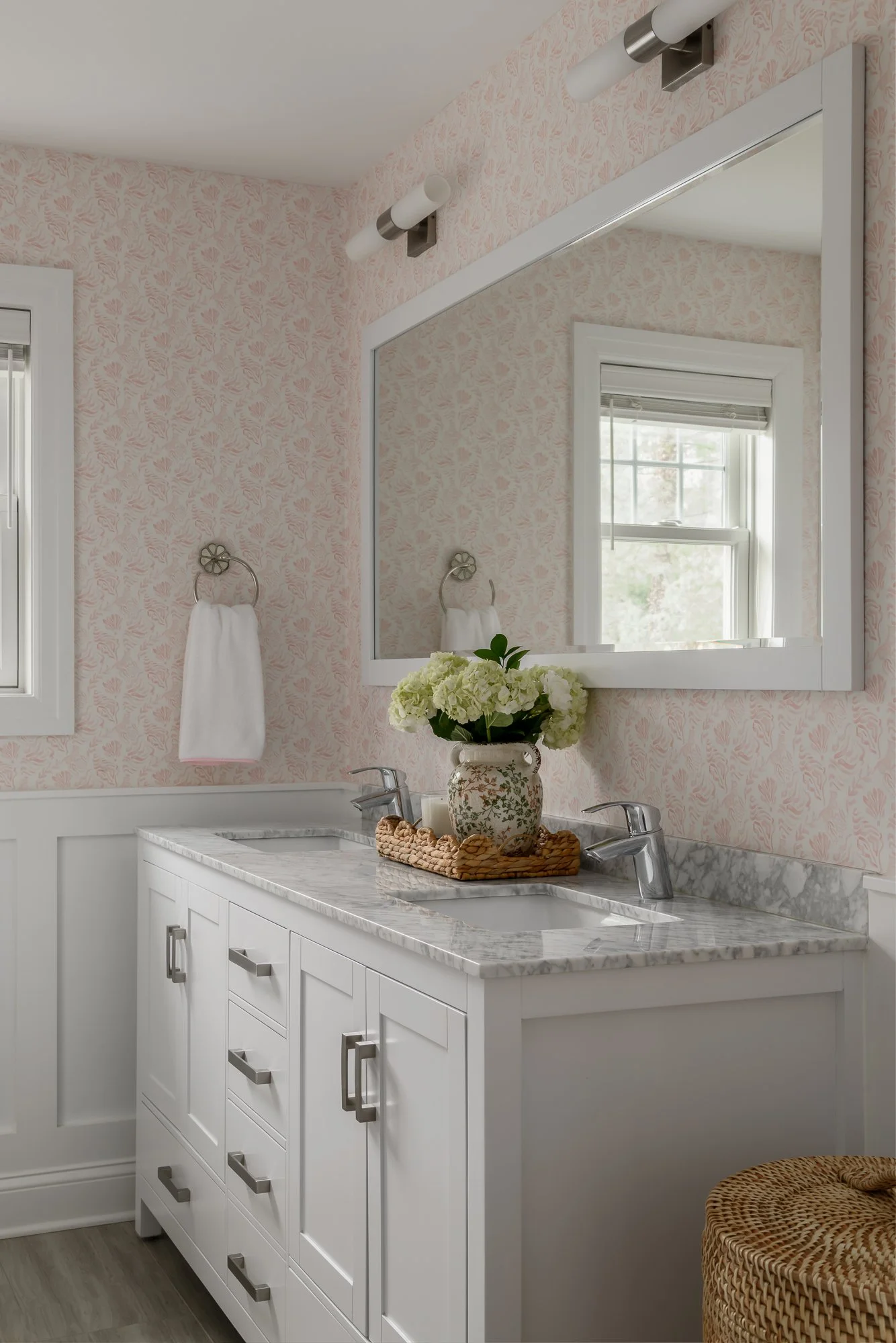 Bathroom vanity with white cabinets, marble countertop, and two sinks. A mirror above the vanity reflects a window, with a flower vase on the counter and pink wallpaper.
