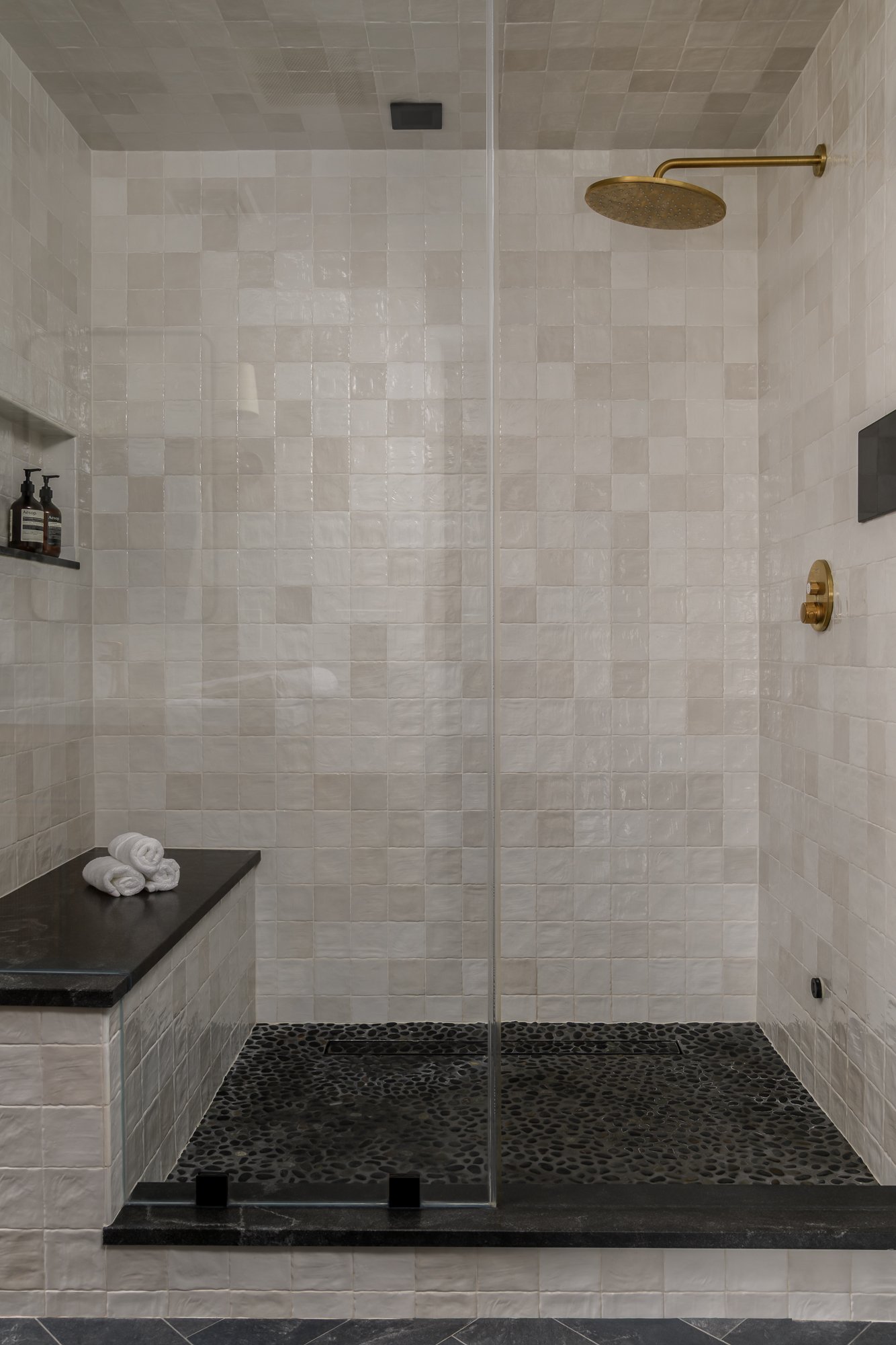 A modern walk-in shower with beige tiled walls, a dark pebble stone floor, a gold showerhead, and a built-in shelf with shampoo bottles. There are white towels folded on a black slab bench.