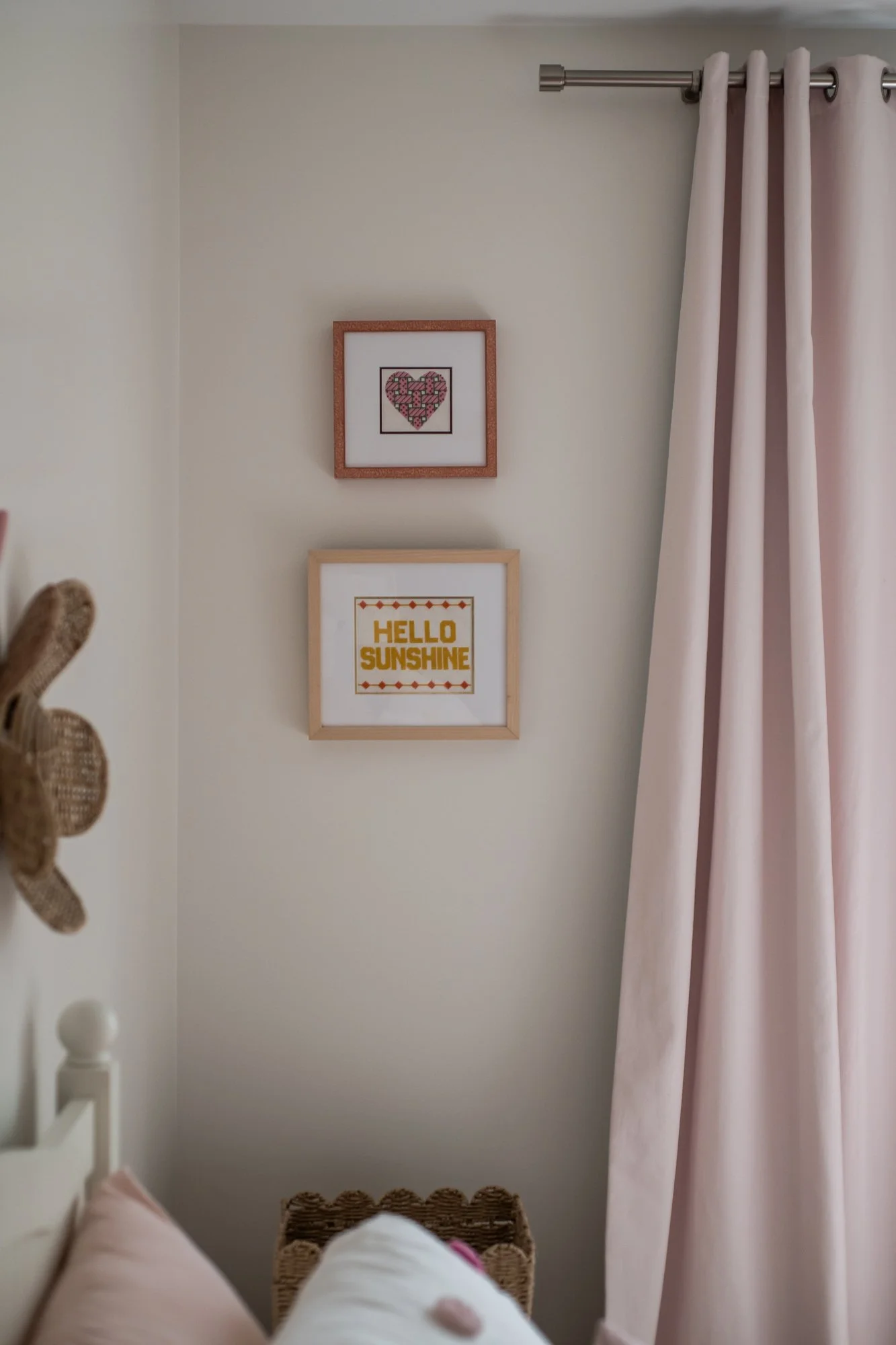 A corner of a room with two framed cross-stitch pictures on the wall. One has a pink heart with a black outline, and the other has yellow text reading "HELLO SUNSHINE" with decorative borders. There are pink curtains hanging on a silver rod to the ri