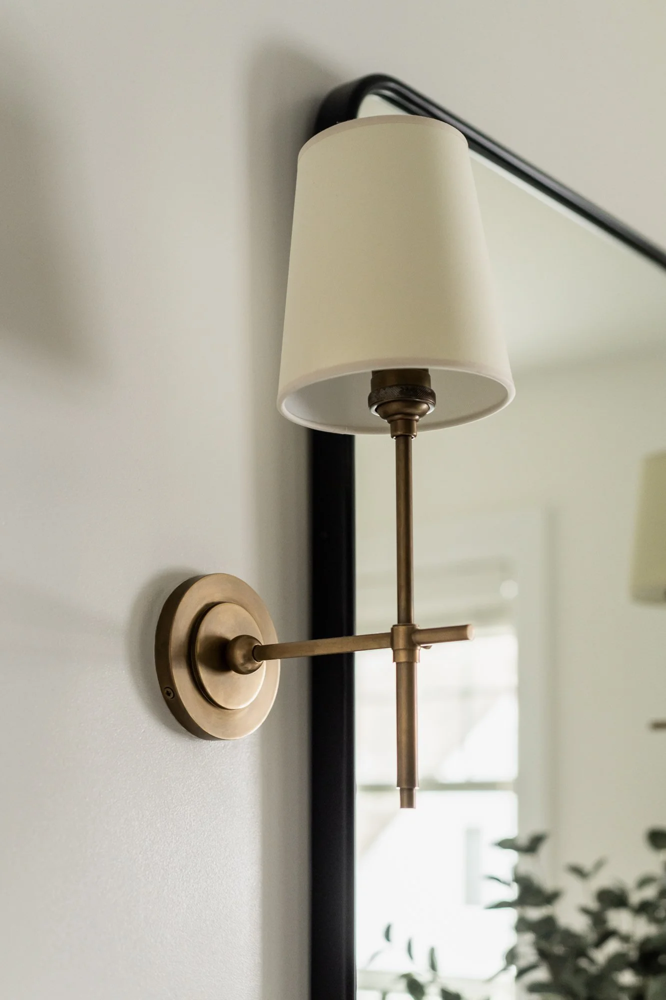 Wall-mounted brass reading lamp with a white lampshade on a white wall near a mirror.