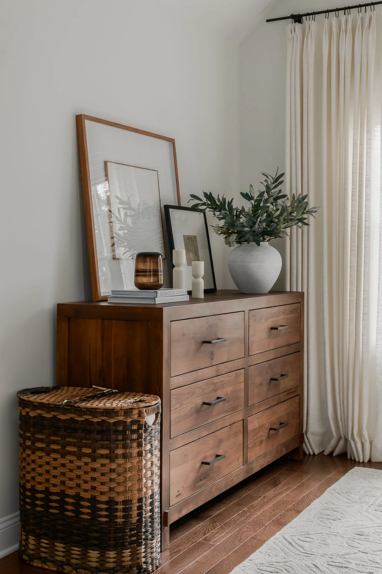 A wooden dresser with six drawers, decorated with framed artwork, a large white vase with greenery, and two white candles. A wicker basket is on the floor beside the dresser, and cream-colored curtains hang beside a window on the right.