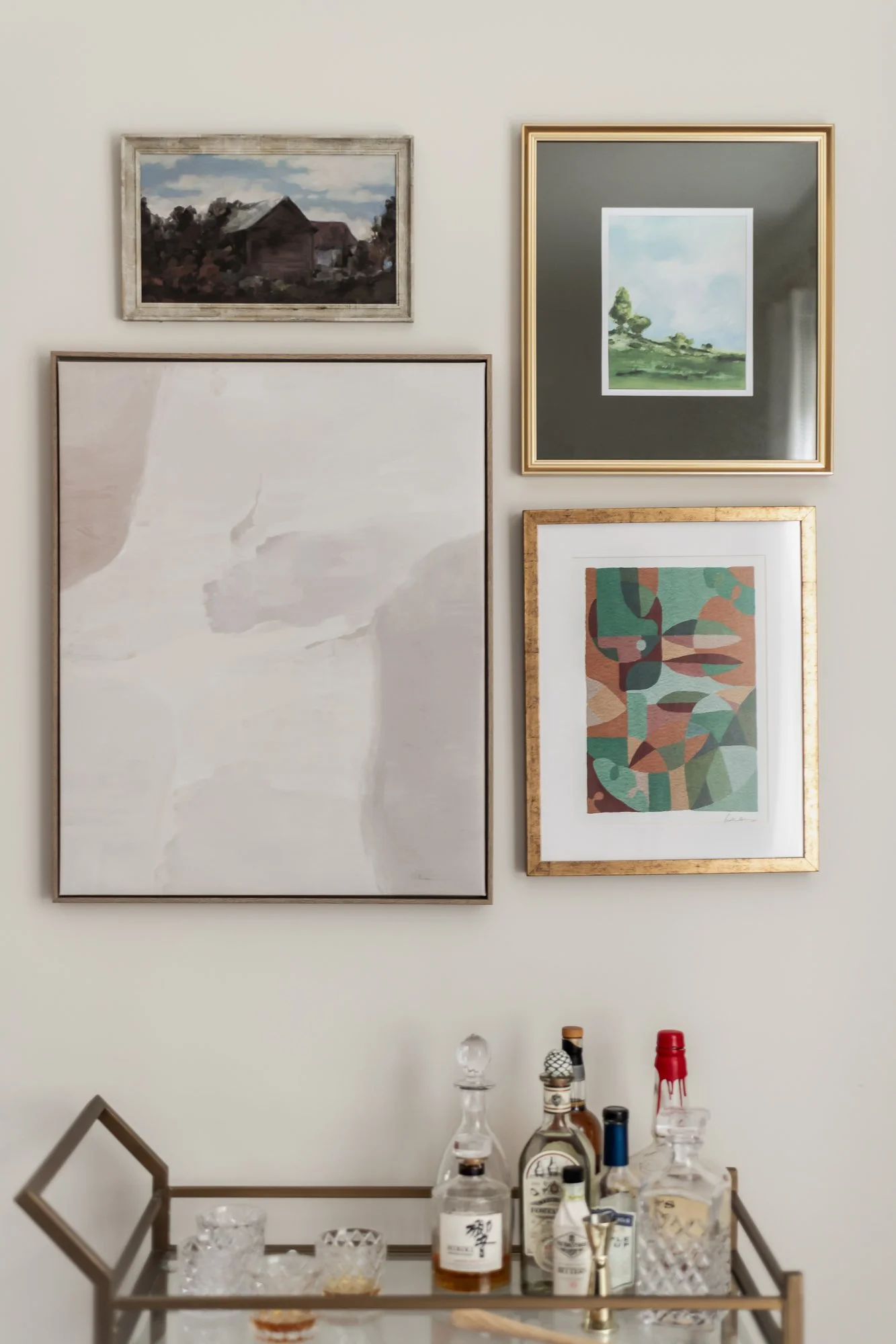 Wall with five framed art pieces in various sizes and styles, a gold and metallic bar cart with bottles of alcohol and glassware in the foreground.