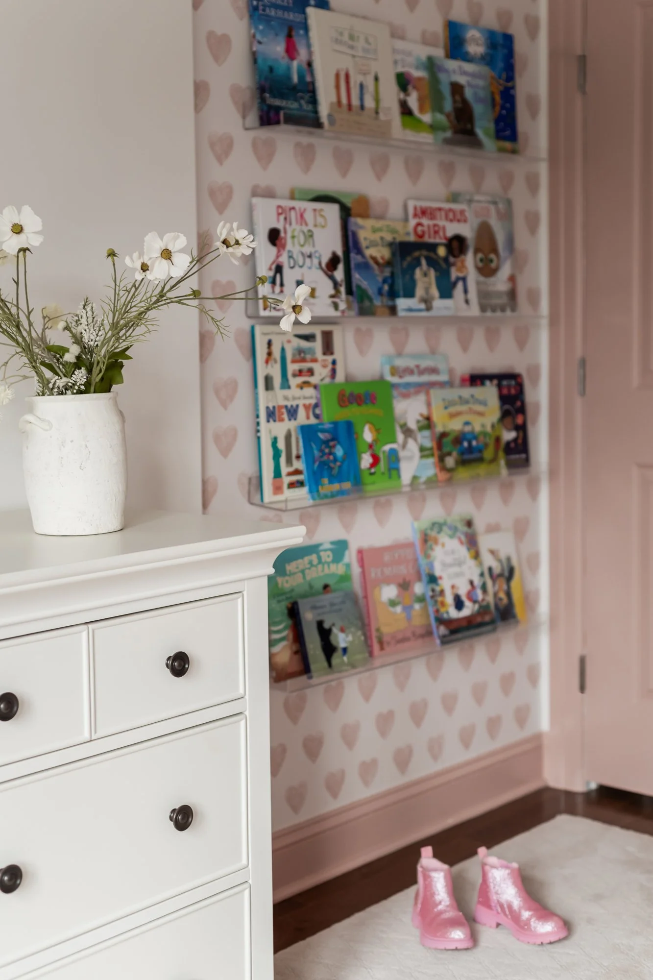 A white dresser with a white vase containing white and green flowers on top. Behind it, a wall covered with pink and beige heart-patterned wallpaper features multiple shelves filled with children's books. On the floor, a white rug and pink rain boots