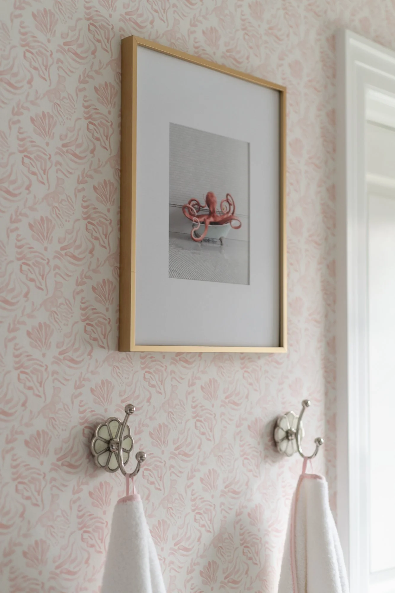 Wall with pink floral wallpaper, framed black and white photo of a pink octopus sculpture, two decorative towel hooks with white towels hanging, near a window with white blinds.