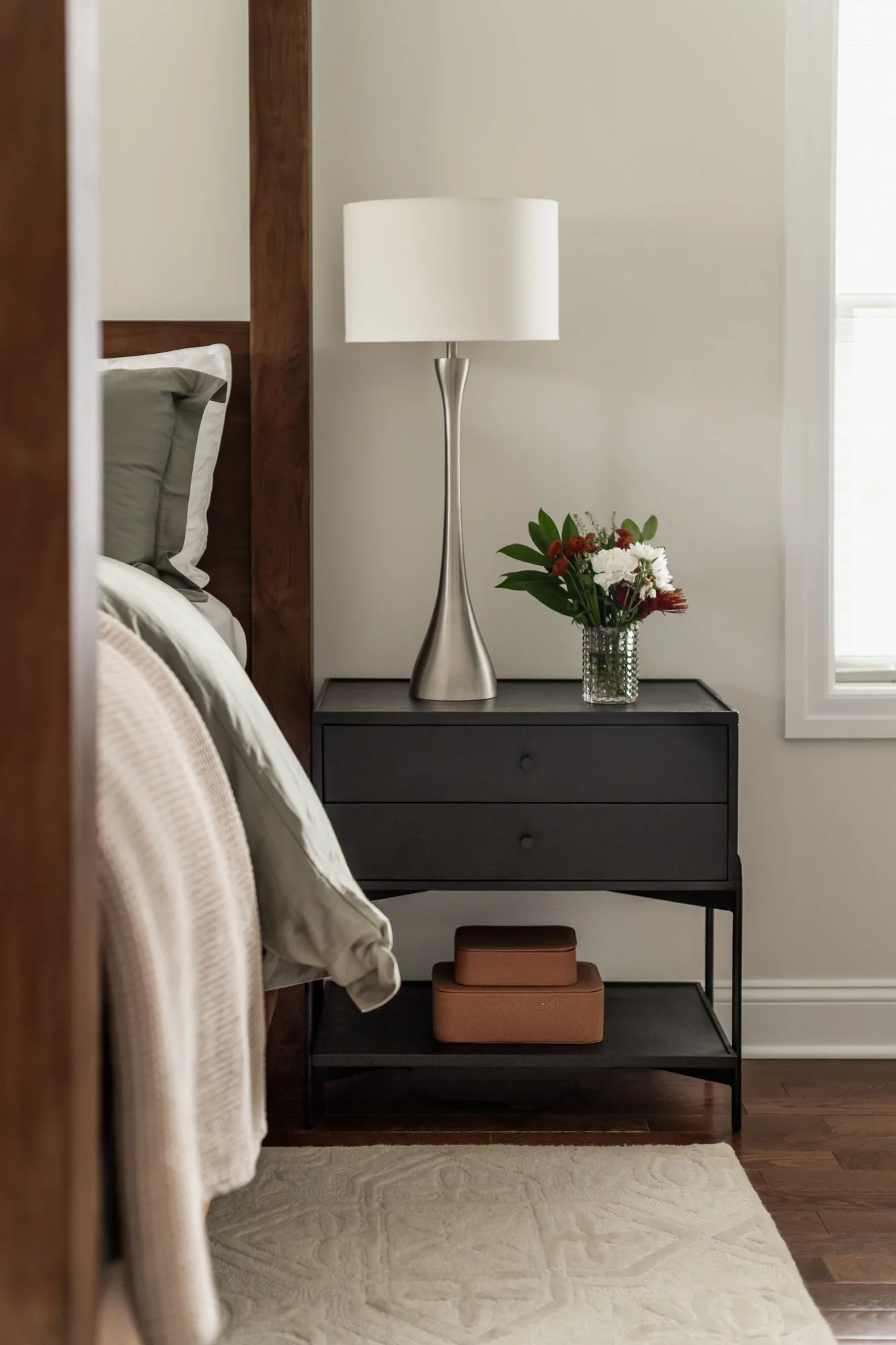 A bedside table with a modern silver lamp, a flower arrangement in a glass vase, and stored boxes underneath, beside a bed with grey and white pillows, in a bedroom with wood flooring and a window.
