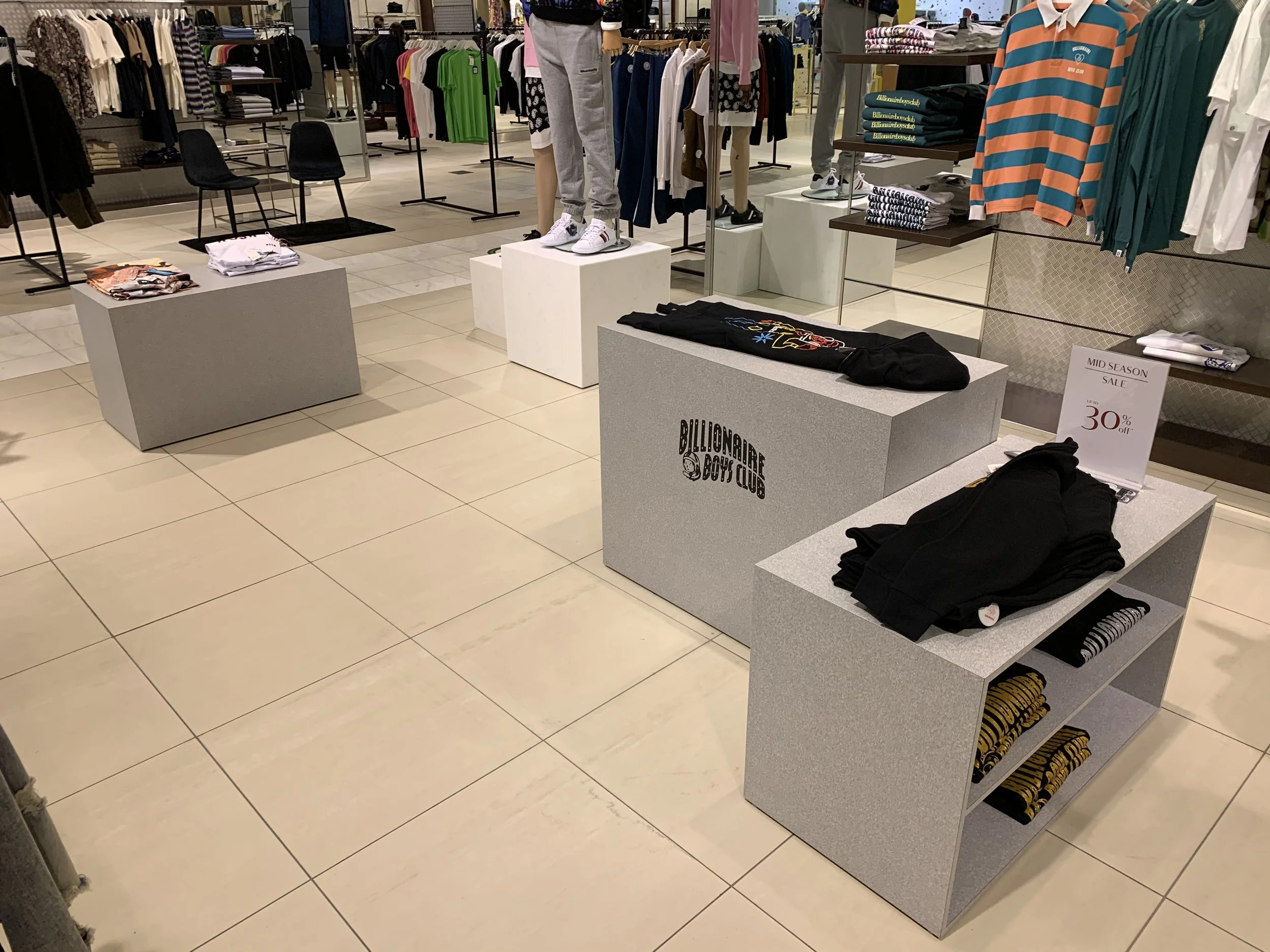 Clothing display in a retail store with folded clothes on tables, mannequins dressed in casual wear, and racks of shirts and pants in the background.