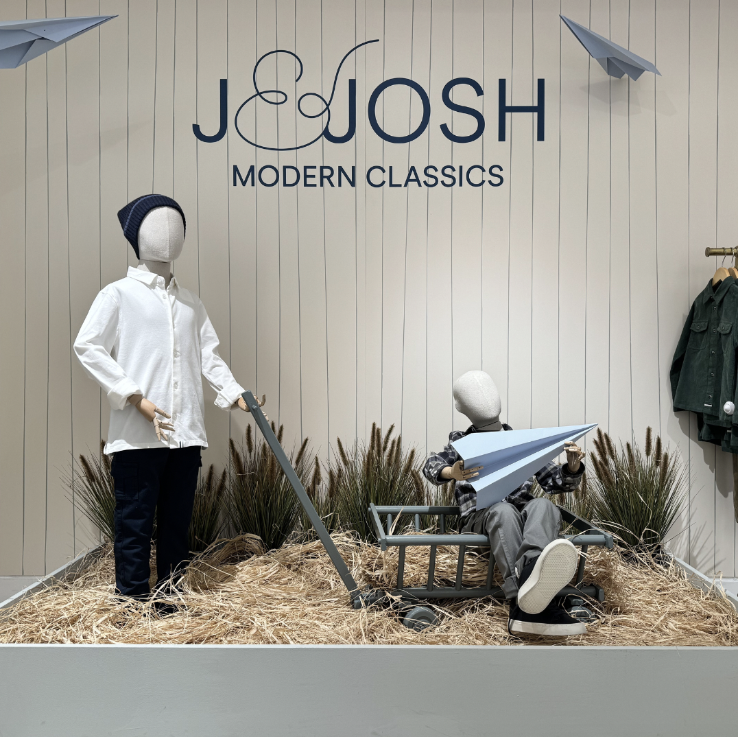 Children's mannequins in a store display with the sign 'JOSH MODERN CLASSICS'; one mannequin stands with a beanie and white shirt, another sits in a cart holding a paper airplane, set on straw with plants behind, decorated with paper airplanes on the