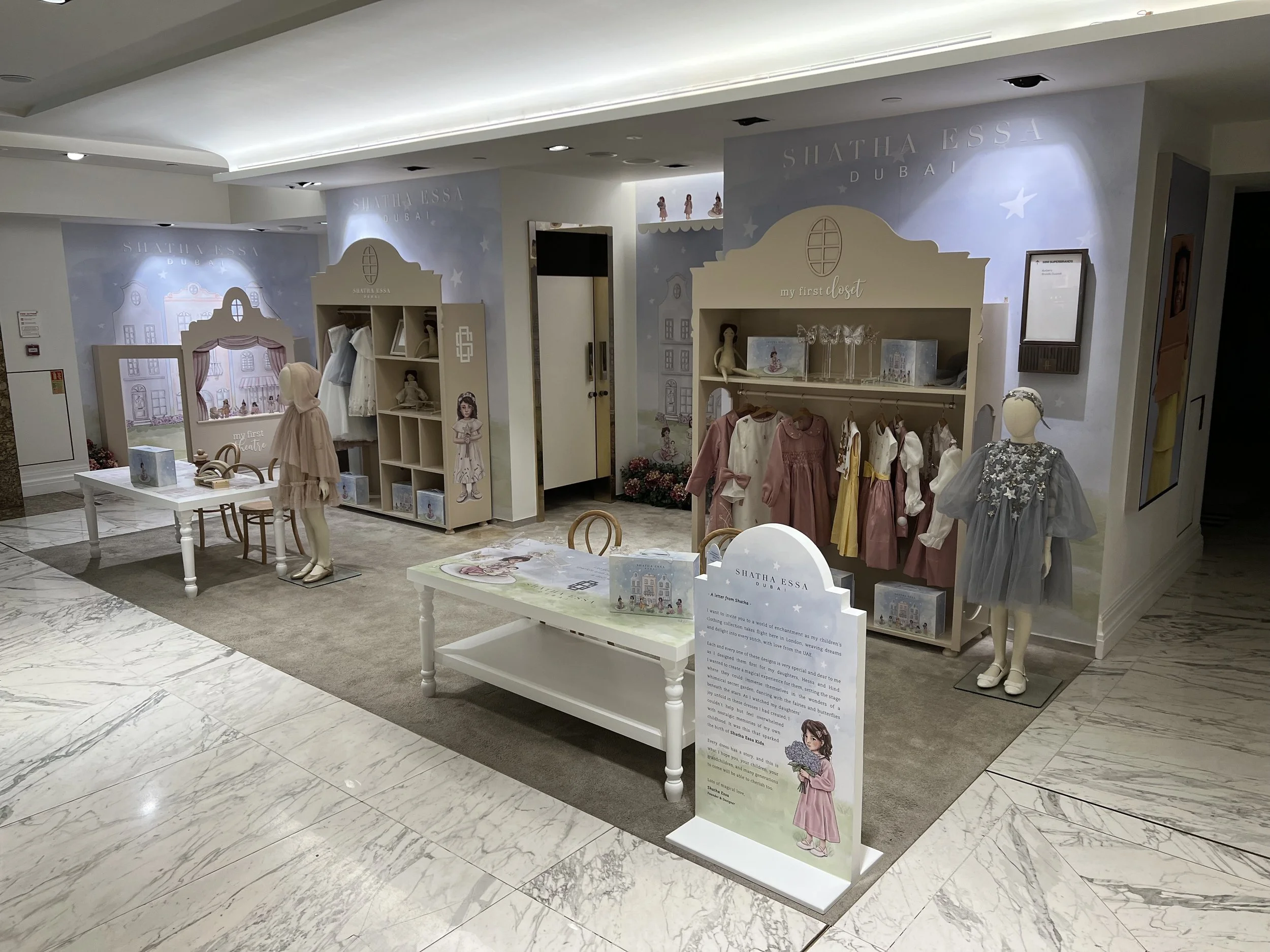 Children's clothing display inside a store with dresses on mannequins and racks, tables with books and decorations, in a well-lit space with marble floors and a painted backdrop of buildings and stars.