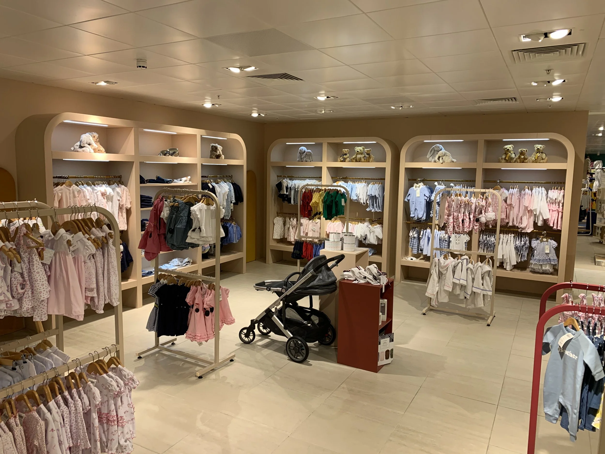 Children's clothing store with beige shelves displaying toys and stuffed animals on top, and racks of children's clothes arranged by size and color. There is a baby stroller in the center.