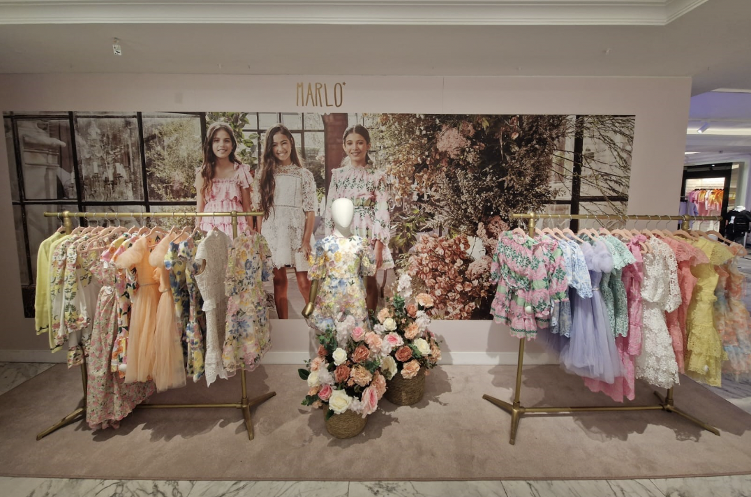 Display of children's dresses on gold clothing racks with a floral backdrop and mannequin in the center, decorated with pink and white flowers.