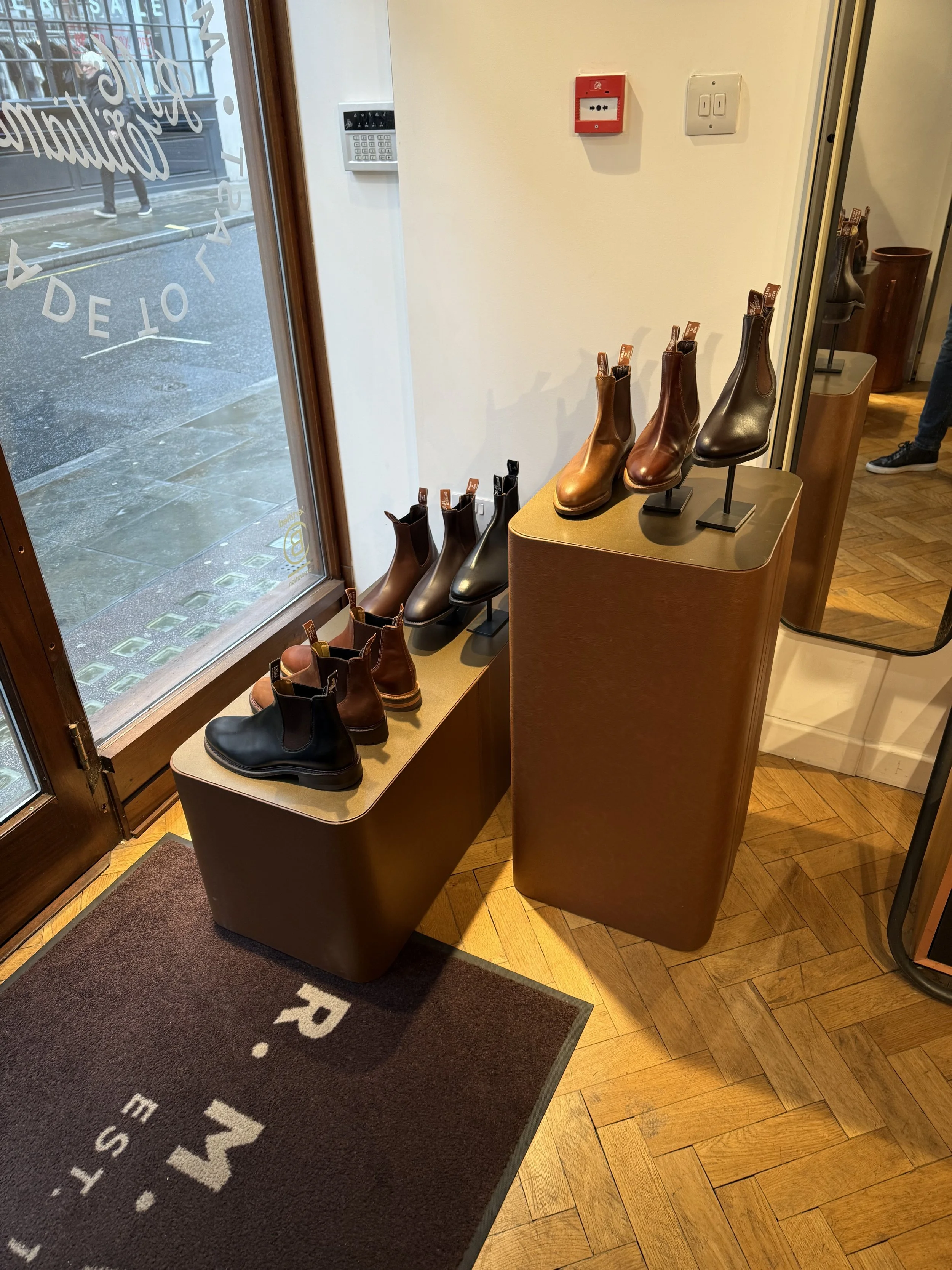 Display of leather boots and shoes arranged on two brown platforms near a glass store entrance, with a sidewalk and pedestrians visible outside through the glass.