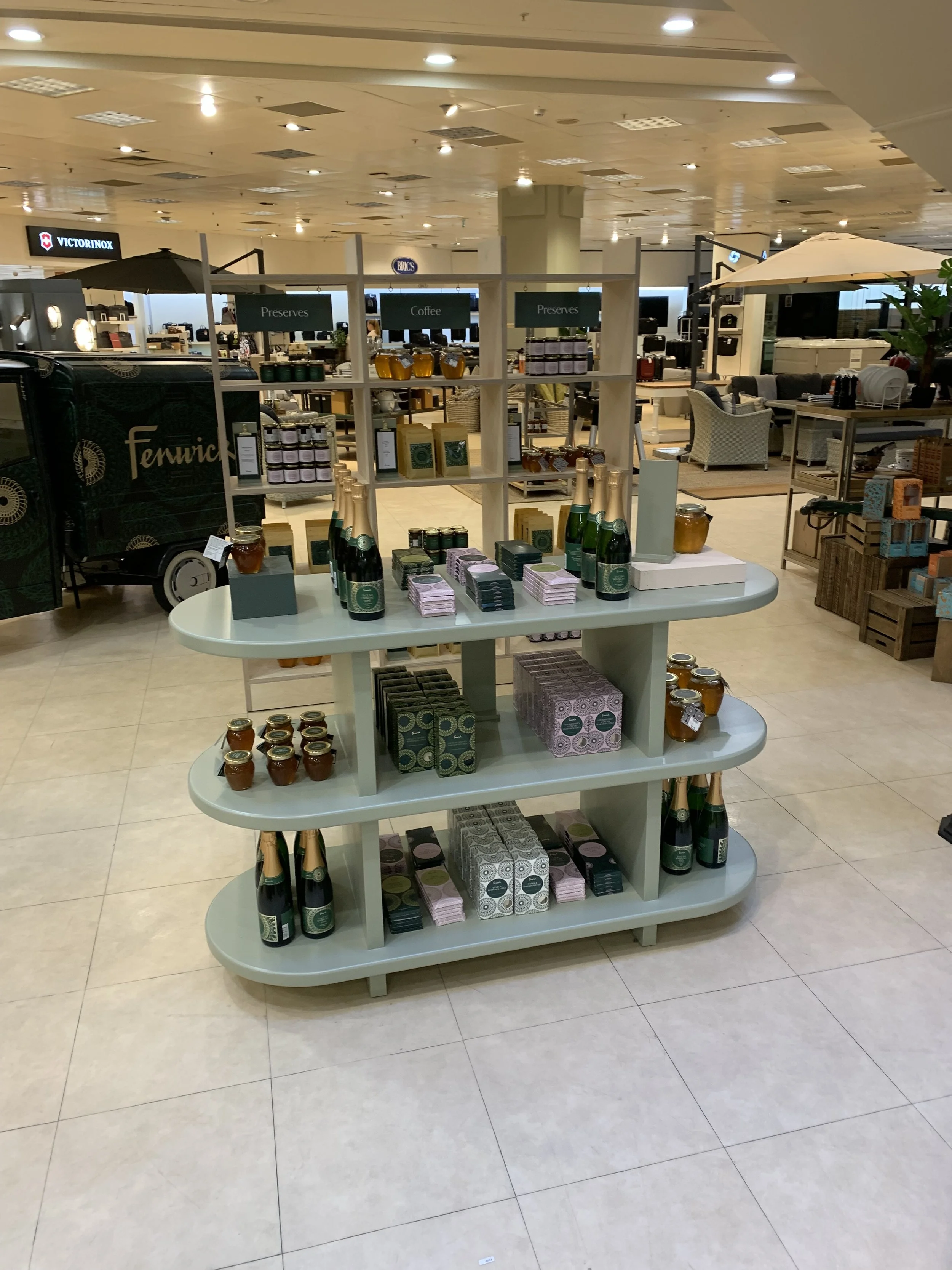 Display of bottled beverages, jars, and boxed products on a round, tiered white shelf in a retail store, with store seating and counters in the background.
