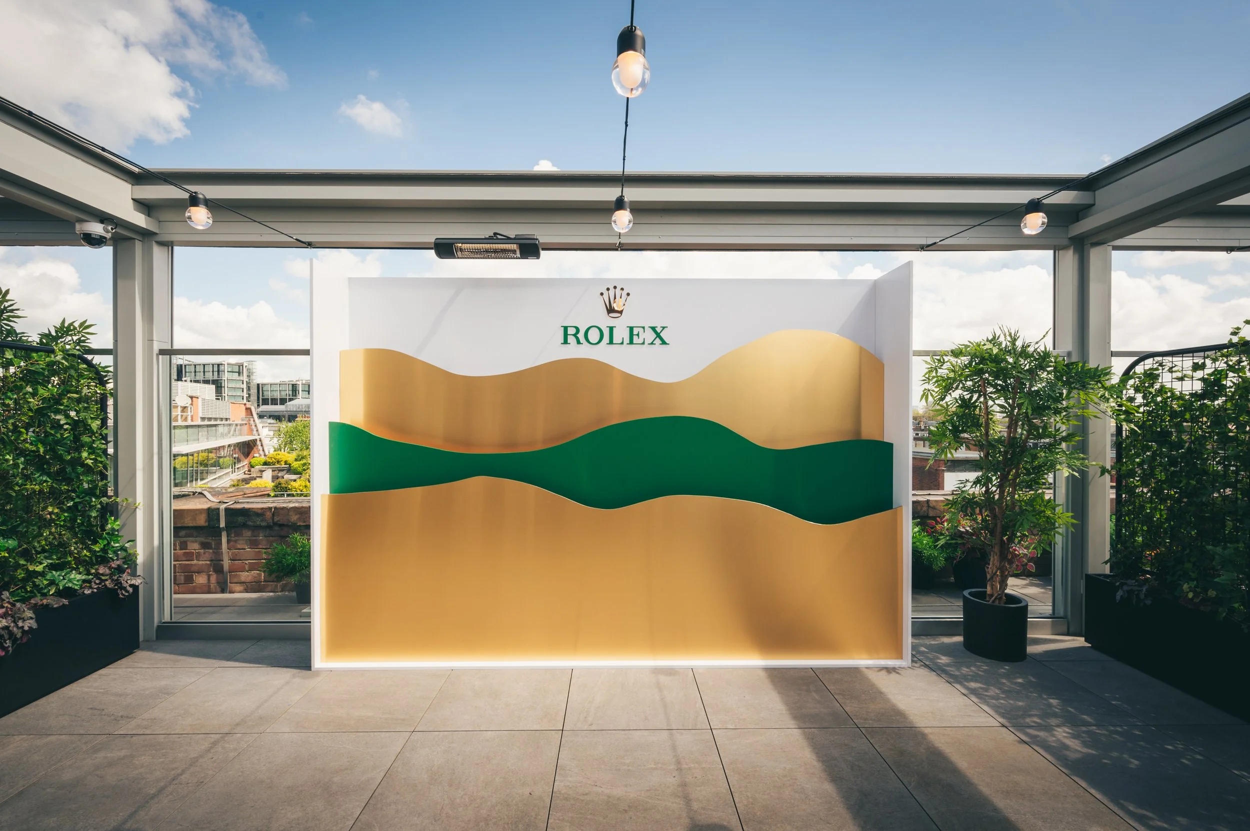 Outdoor balcony with potted plants, a white wall with a green and gold abstract design and Rolex logo, and a clear blue sky