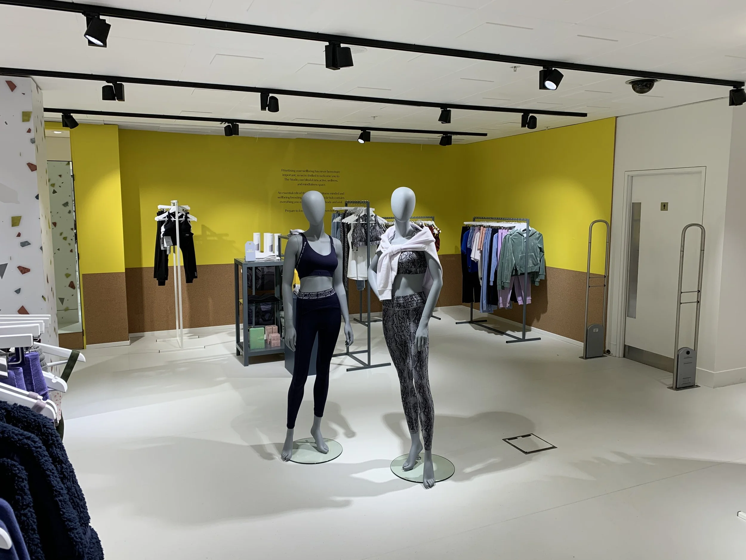 Mannequins dressed in workout clothes inside a retail clothing store with racks of apparel and a bright yellow wall in the background.