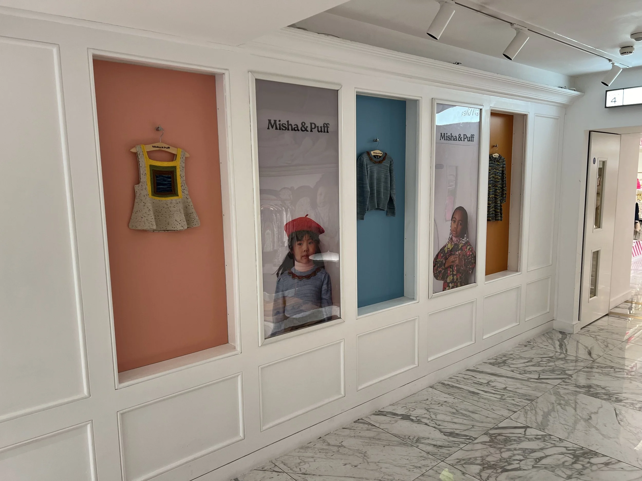 Display window of a children's clothing store with colorful backgrounds and mannequins wearing children's clothing, including a dress, sweater, and jacket, with promotional images and