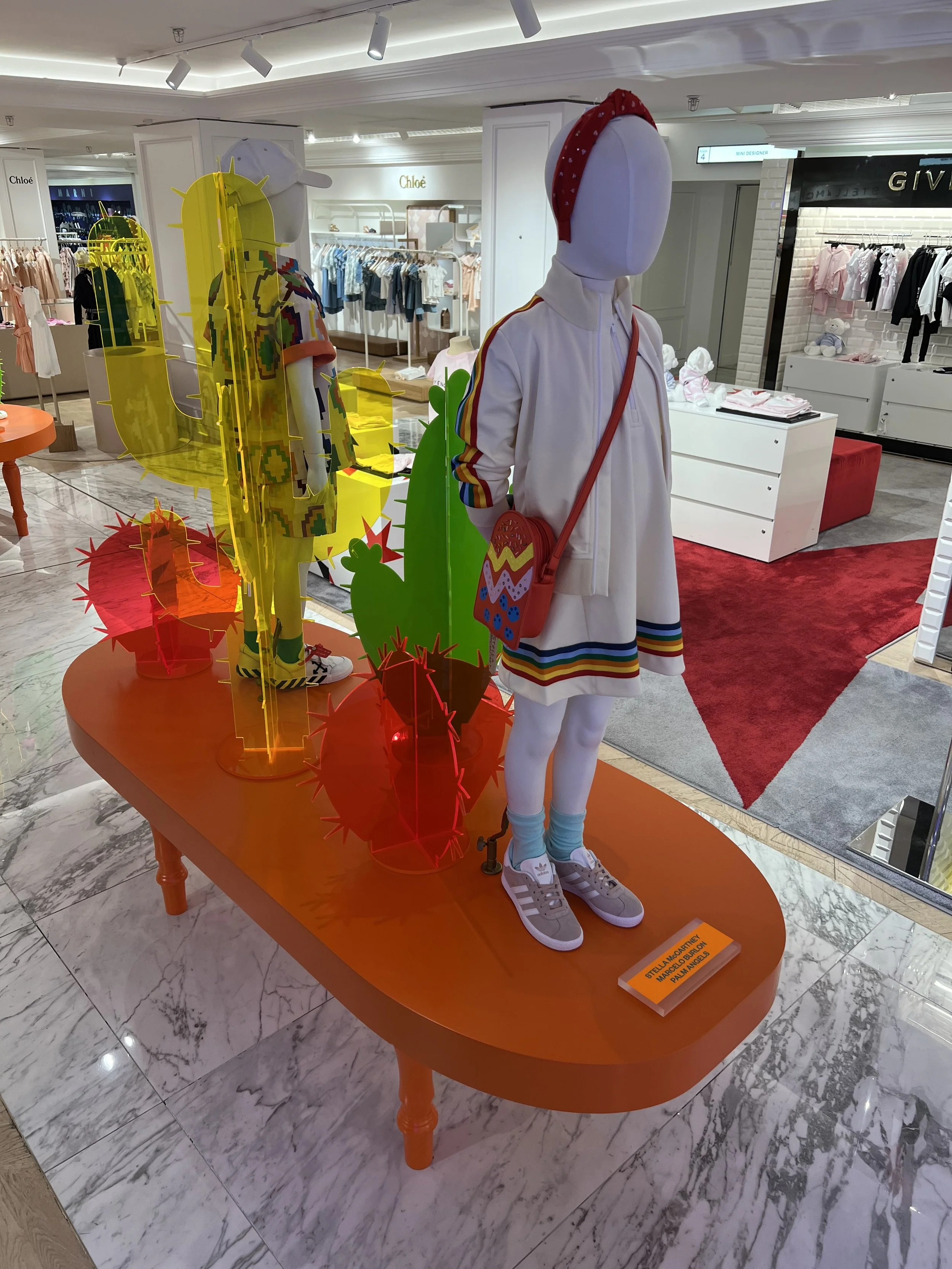 Mannequin dressed in a white athletic jacket and skirt with rainbow stripes, wearing sneakers and light blue socks, carrying a colorful embroidered bag, standing on an orange display table with red cactus-shaped decorations. In the background, store