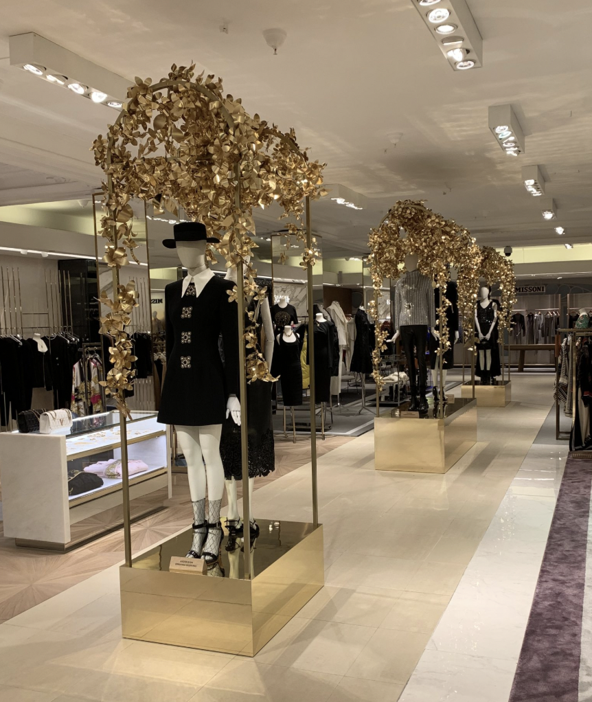 Fashion mannequins dressed in black outfits with gold decorative arches and floral arrangements in a clothing store.