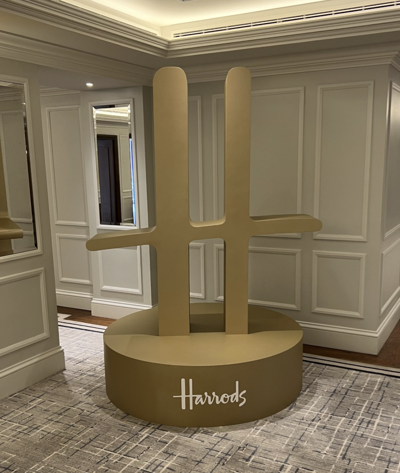 A large, gold-colored hashtag symbol sculpture with the word 'Harrods' written on its rounded base, placed in an elegant interior space with white paneled walls and a patterned carpet.