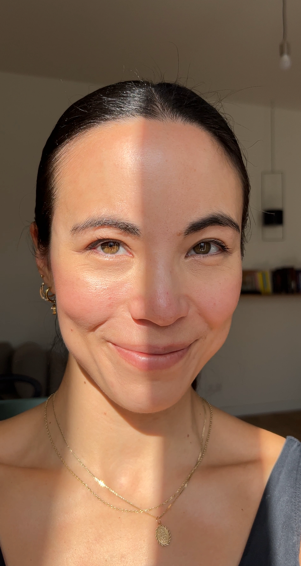 A smiling woman with dark hair pulled back, wearing gold hoop earrings and layered necklaces, in sunlight indoors.