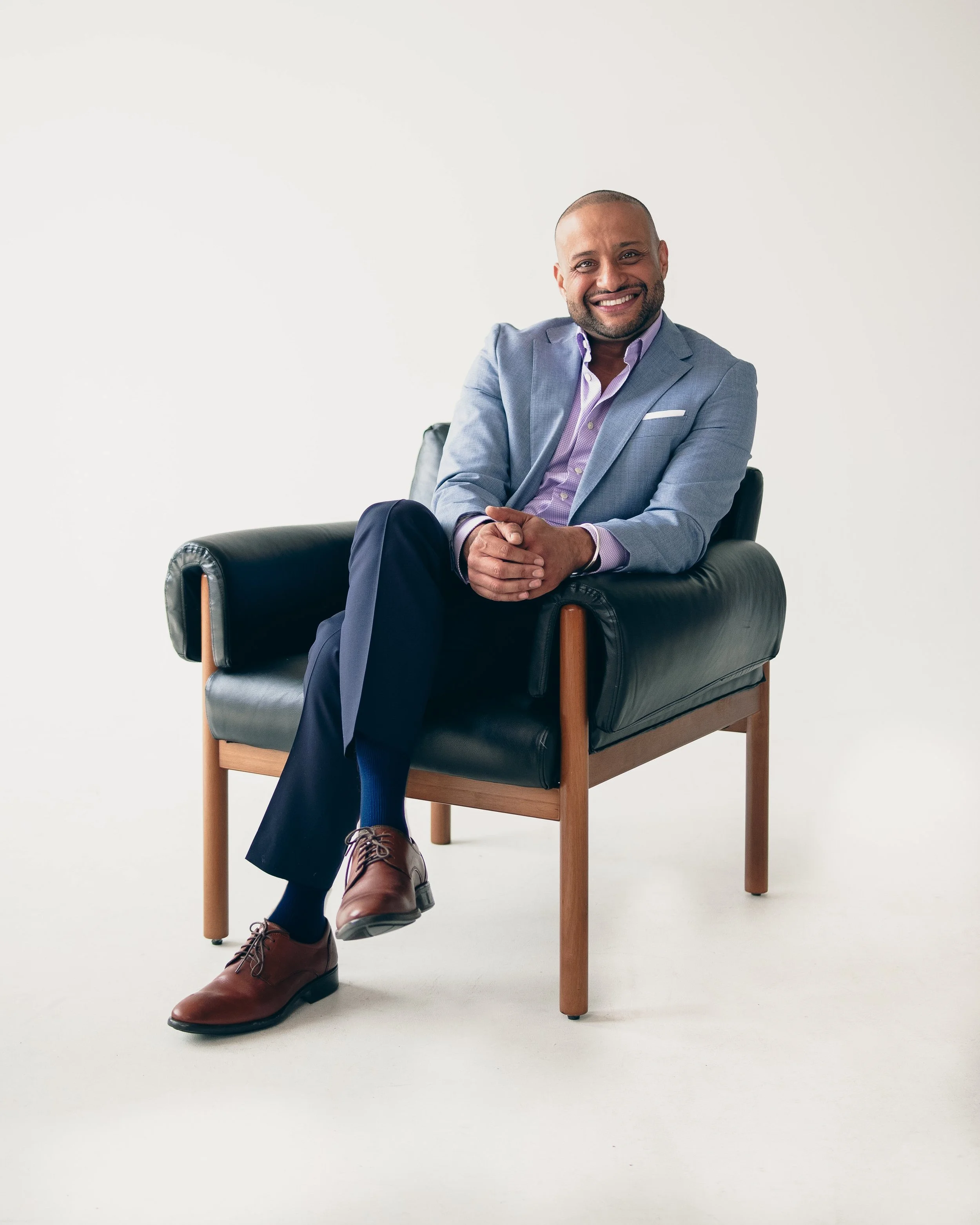 A man in a light gray blazer and navy pants sitting on a black leather armchair with a light wood frame, smiling at the camera against a plain white background.