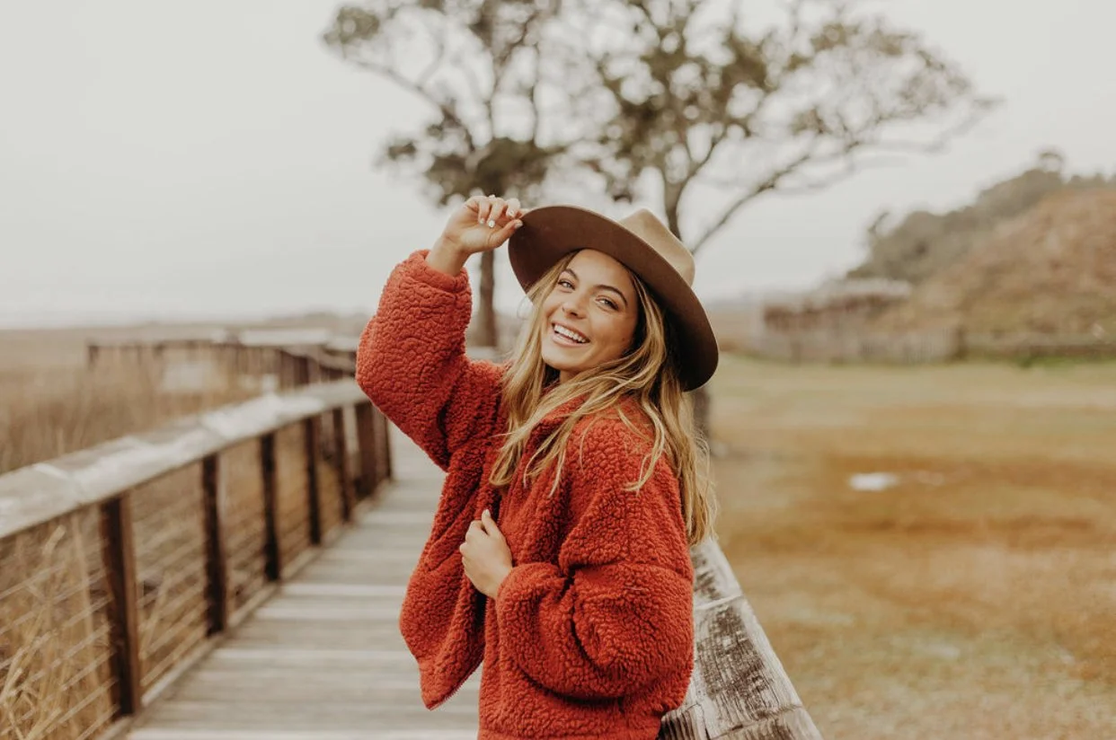 Young woman in orange furry jacket and brown hat smiling on wooden boardwalk in outdoor setting with trees and grass