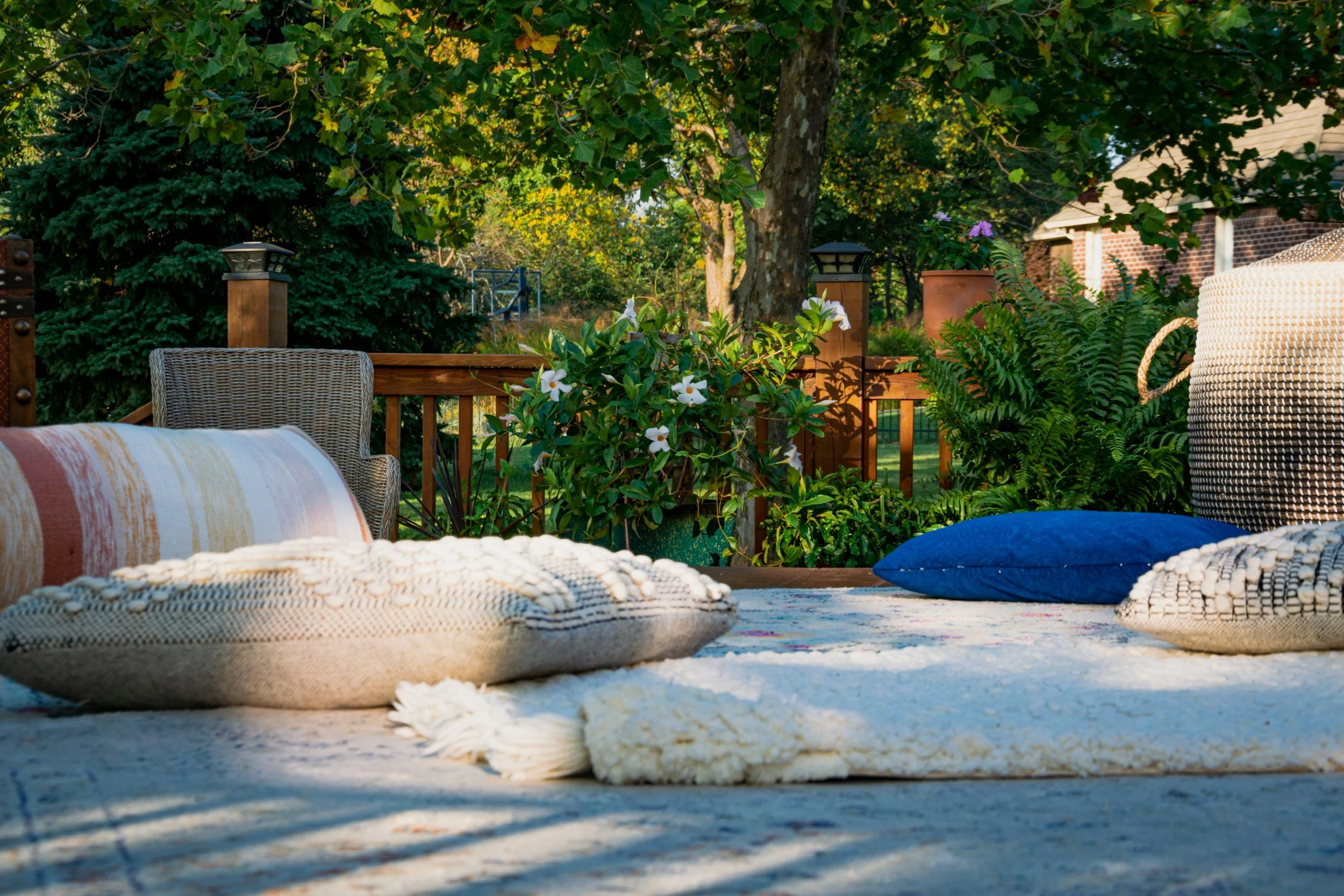 Outdoor patio with colorful cushions, blankets, and pillows on a rug, surrounded by greenery and trees.