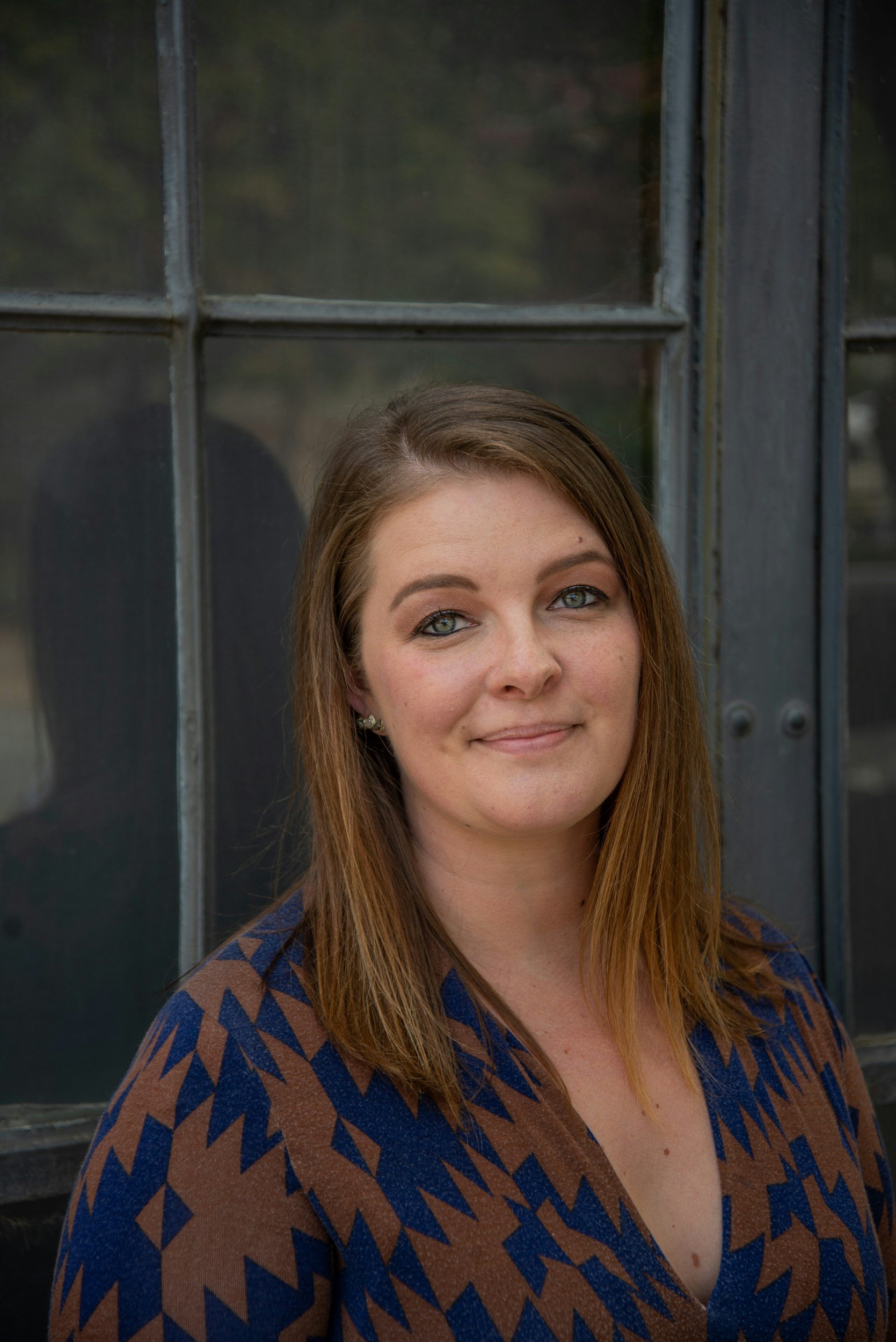A woman with long brown hair and blue eyes standing in front of a window with black wooden framing.