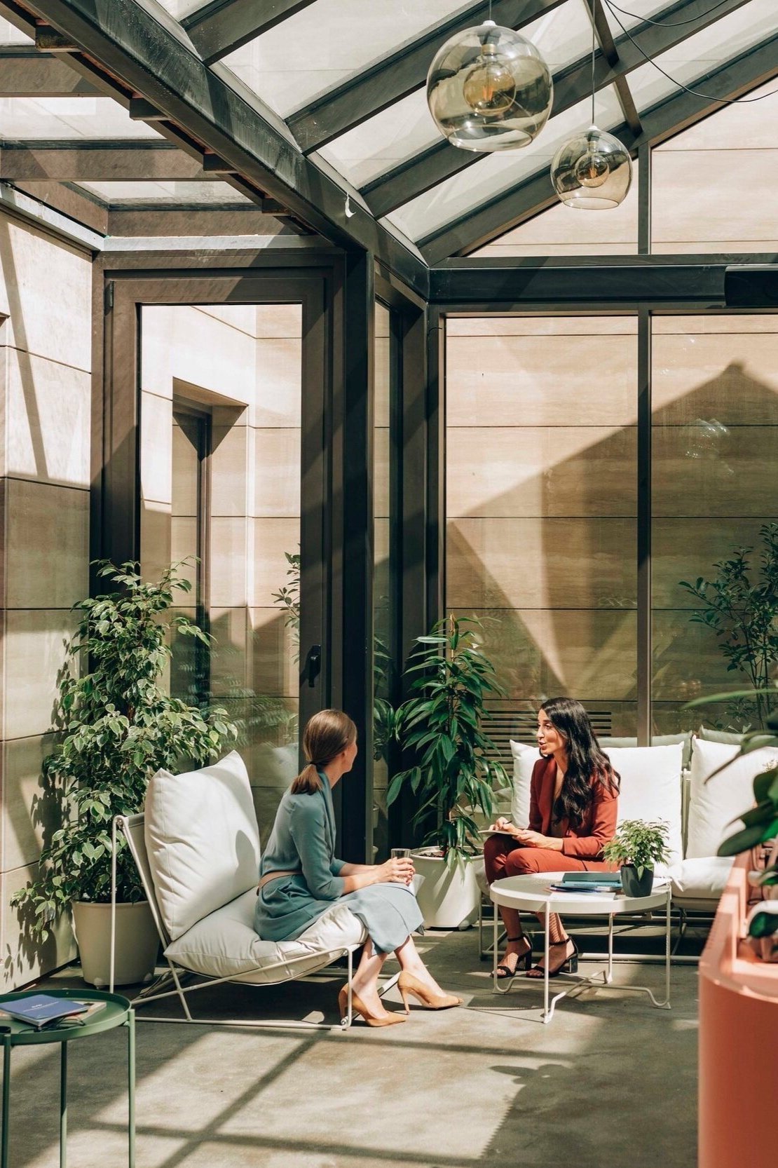 Two women sitting and talking inside a glass conservatory with plants, modern furniture, and pendant lighting.