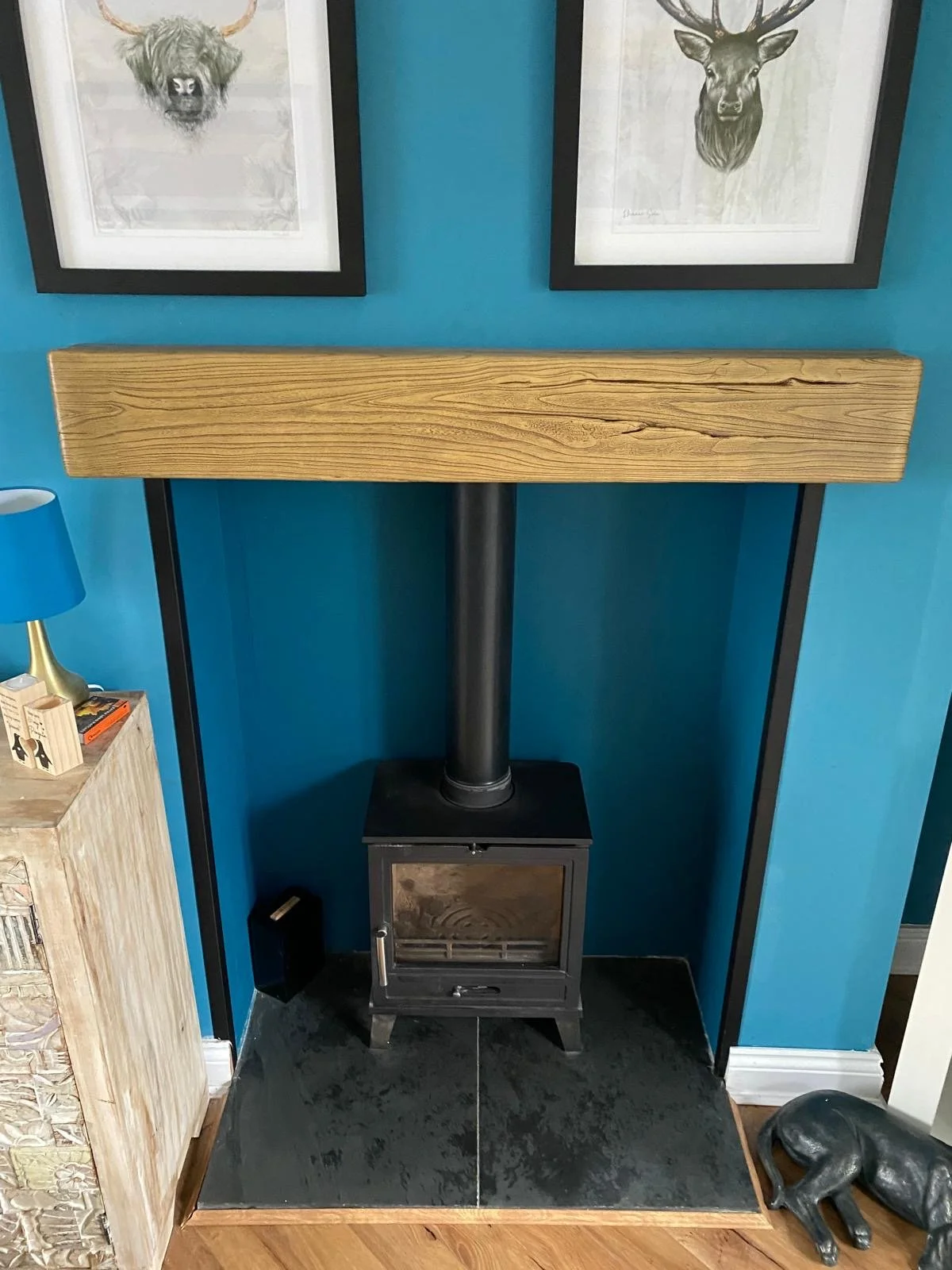 A wood stove with a black pipe, in front of a blue wall, with two framed animal art prints hanging above, and a small side table with a lamp and books on the left. A decorative animal sculpture is on the floor to the right.