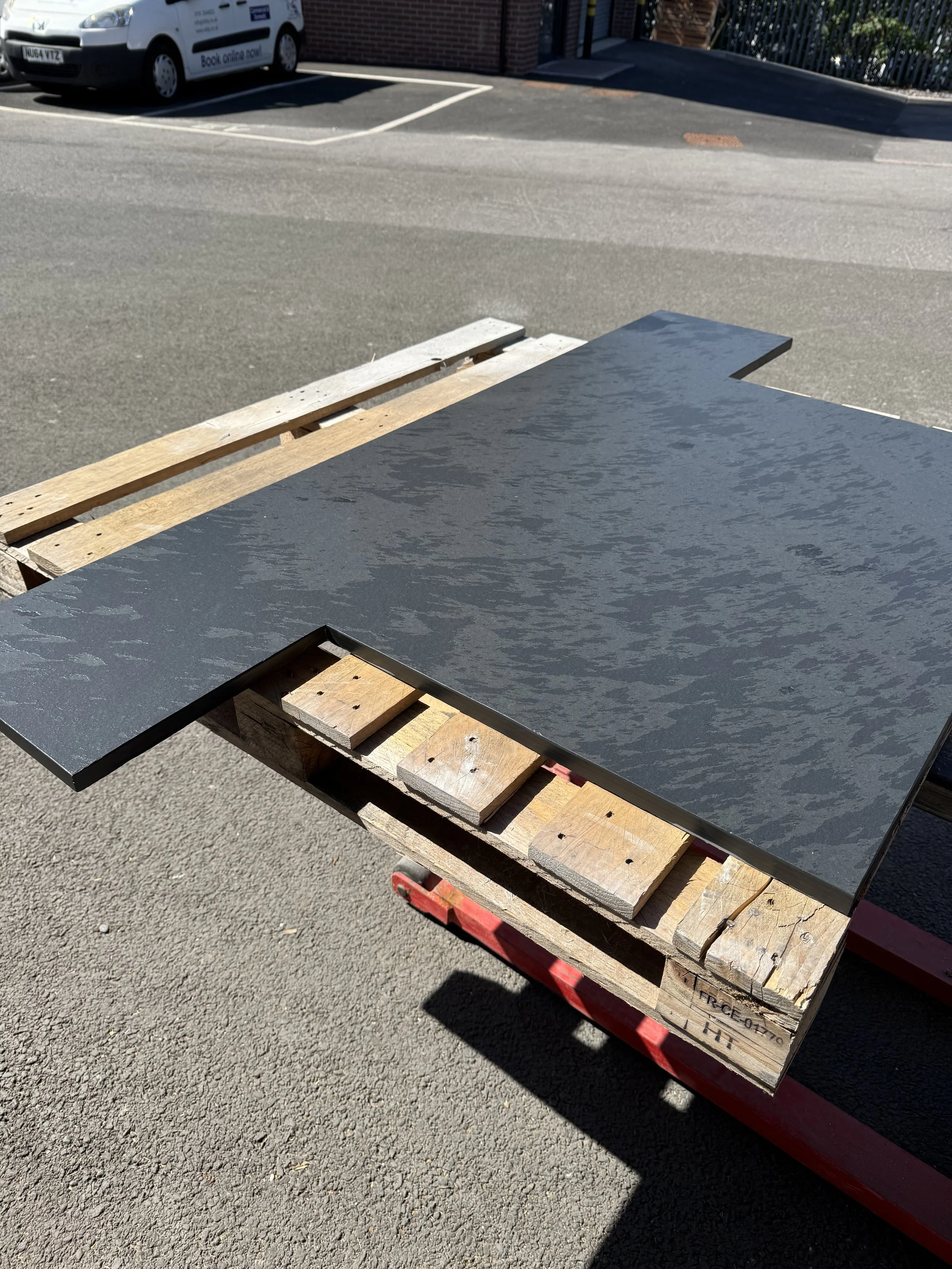 A black countertop with a textured surface resting on a wooden pallet outdoors, with a parking lot and white van in the background.