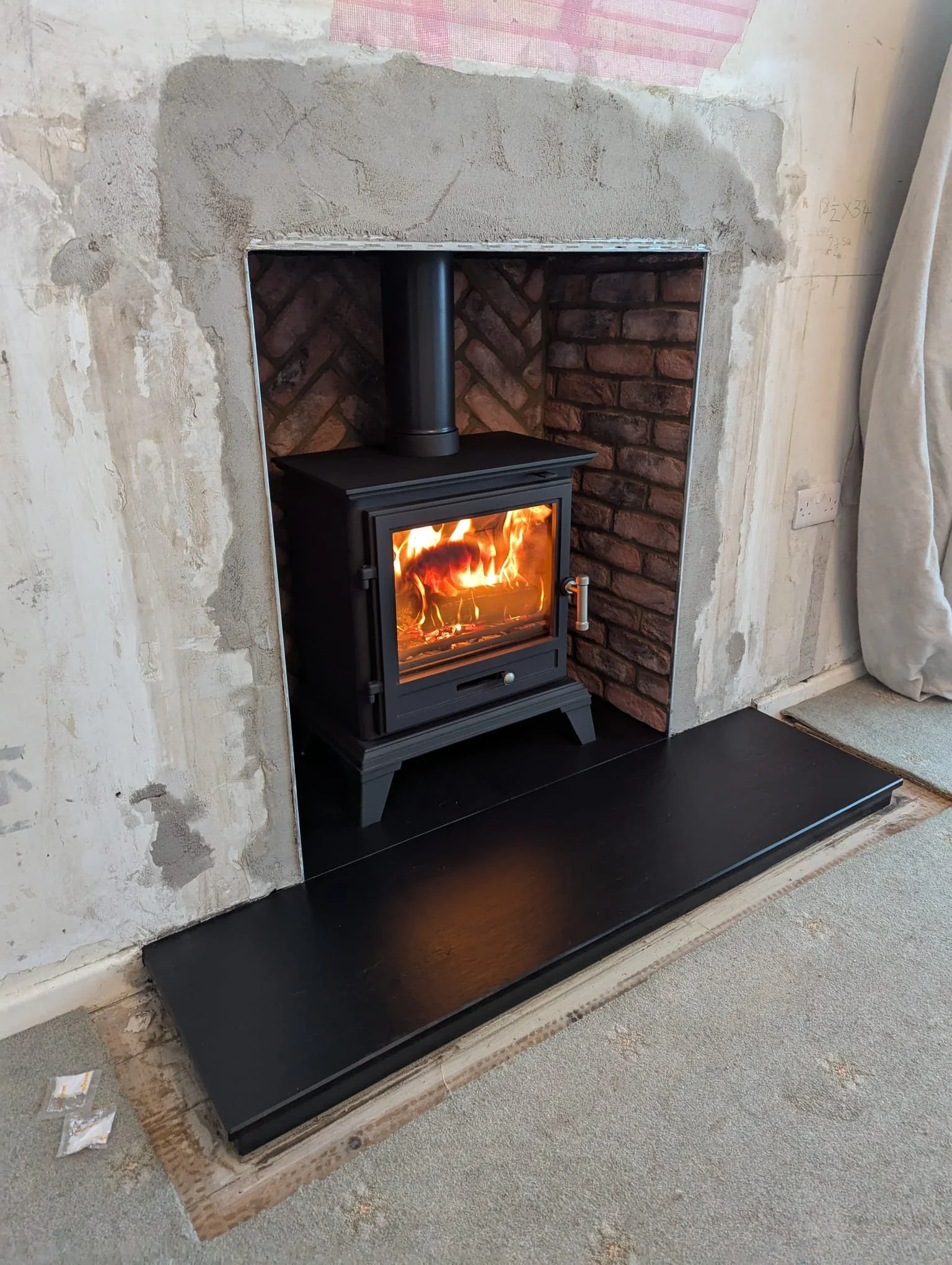 A black wood stove with a glass door showing a fire inside, installed in a brick-lined alcove with a black hearth in a room under renovation.