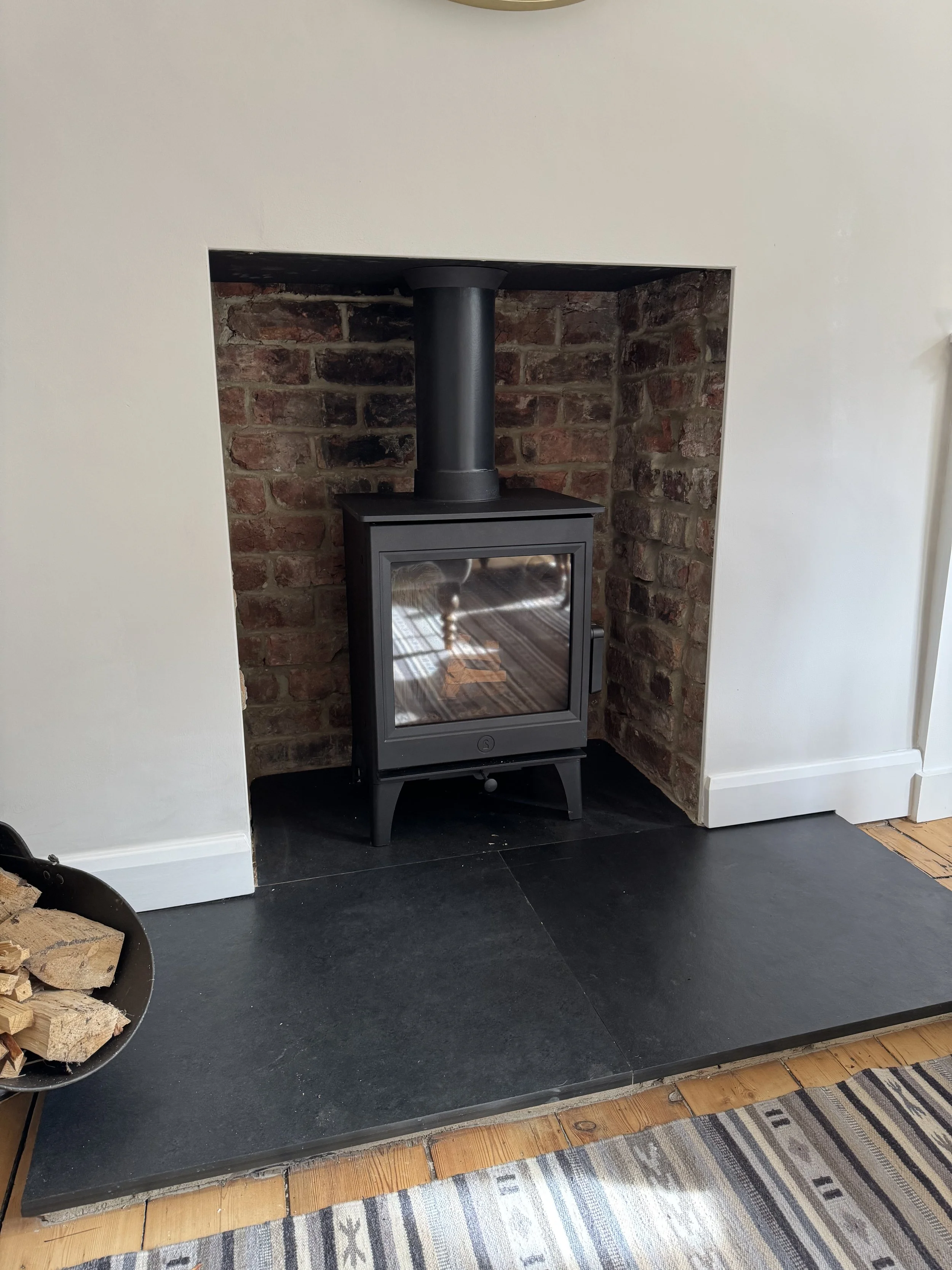 Wood-burning stove in a fireplace with a black chimney pipe and brick interior, on a black slate hearth with a striped rug nearby.