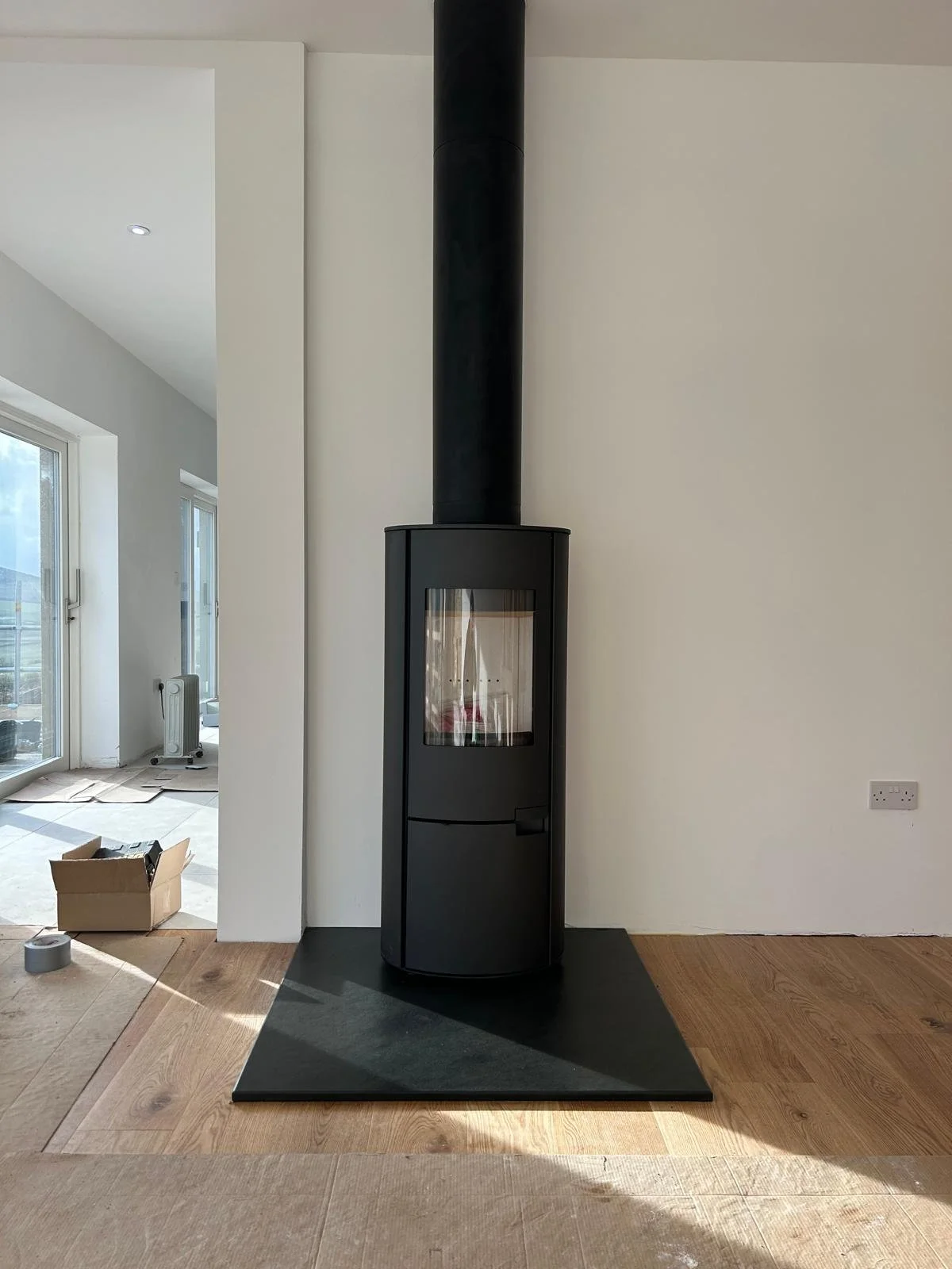 A modern black wood stove on a black hearth pad in a living room with light wood floors and white walls.