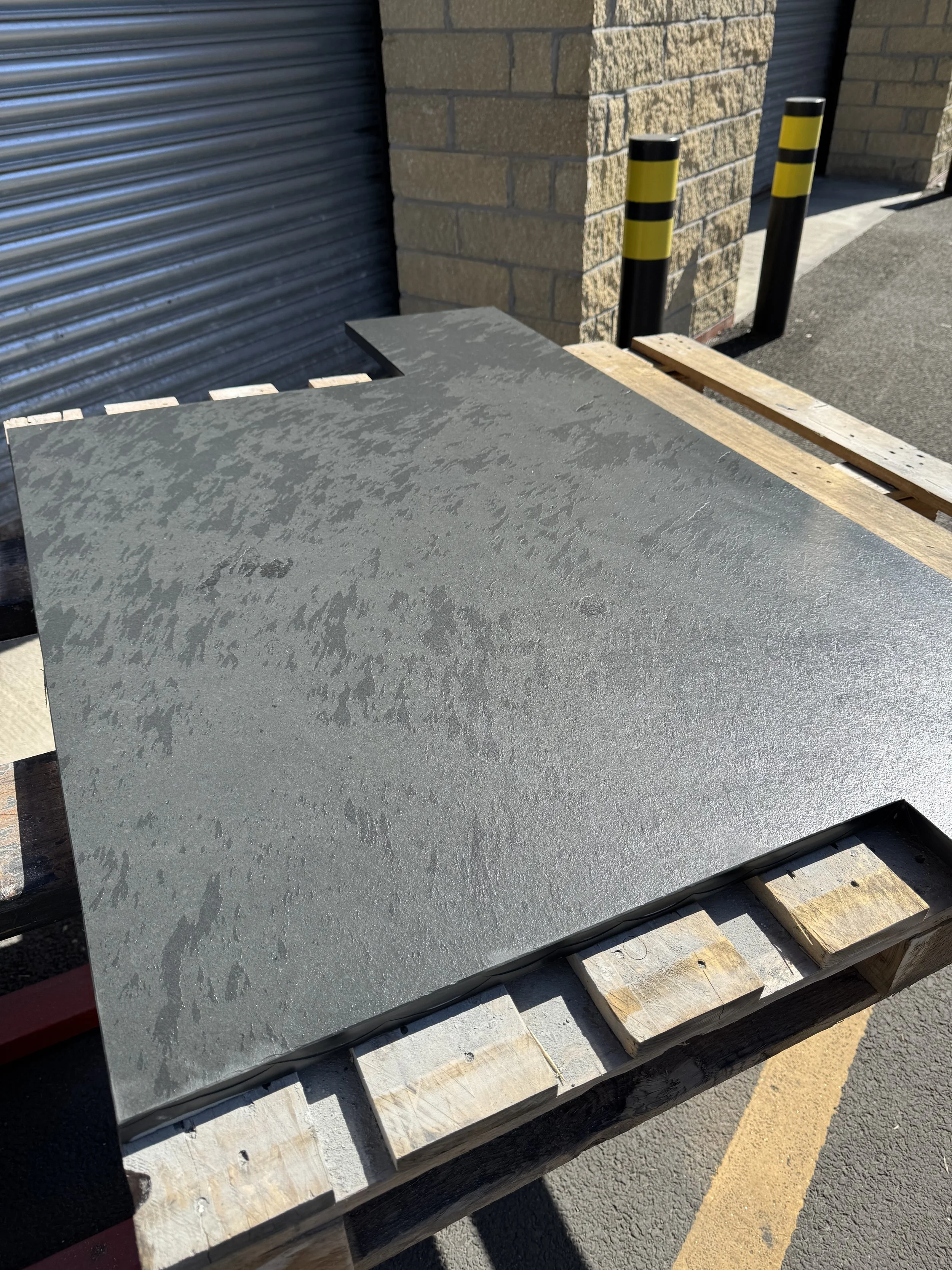 A concrete slab placed on a wooden pallet outside, next to a brick wall and black bollards with yellow stripes.