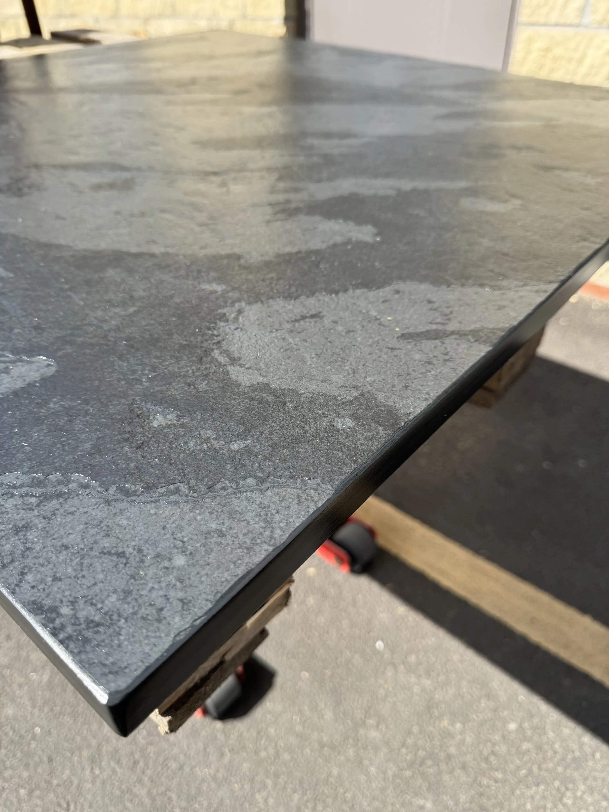 Close-up of a black, textured tabletop with some water spots, supported by a wooden frame with wheels underneath, set outdoors on pavement.