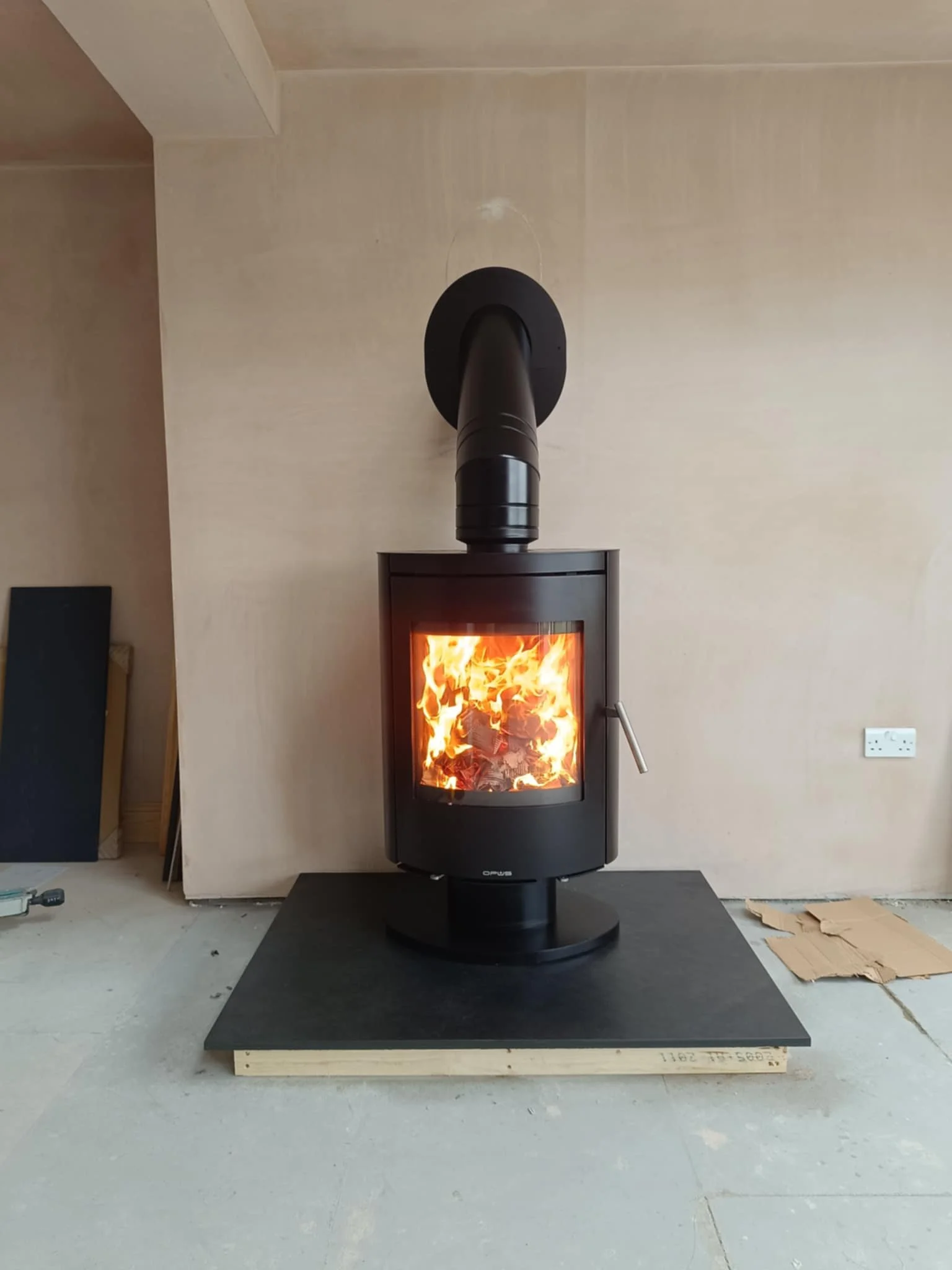 A black wood-burning stove with a glass door showing burning firewood, placed on a black hearth pad against a beige wall.