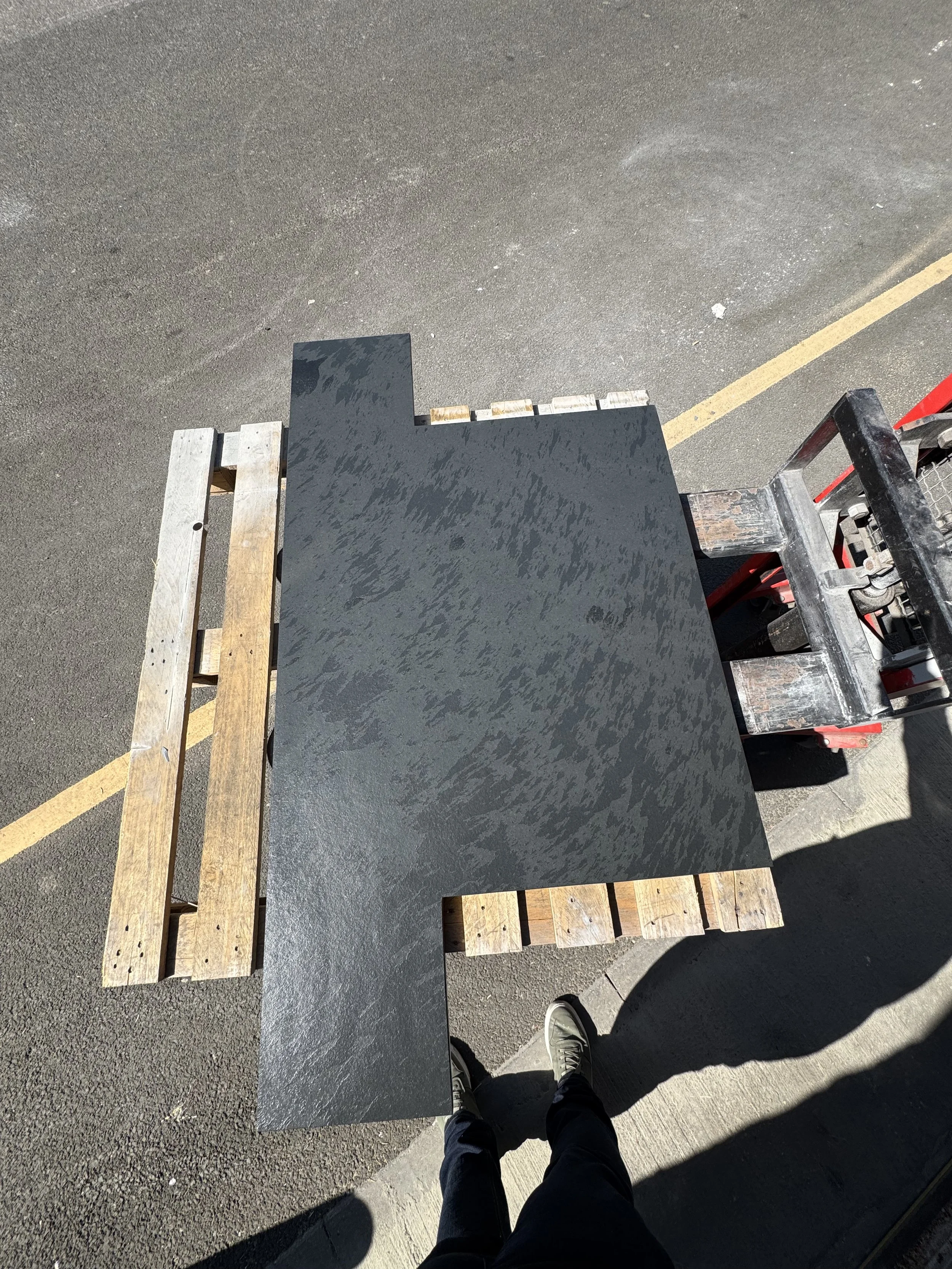 A black, L-shaped granite countertop resting on wooden pallets and a forklift on an outdoor asphalt surface.