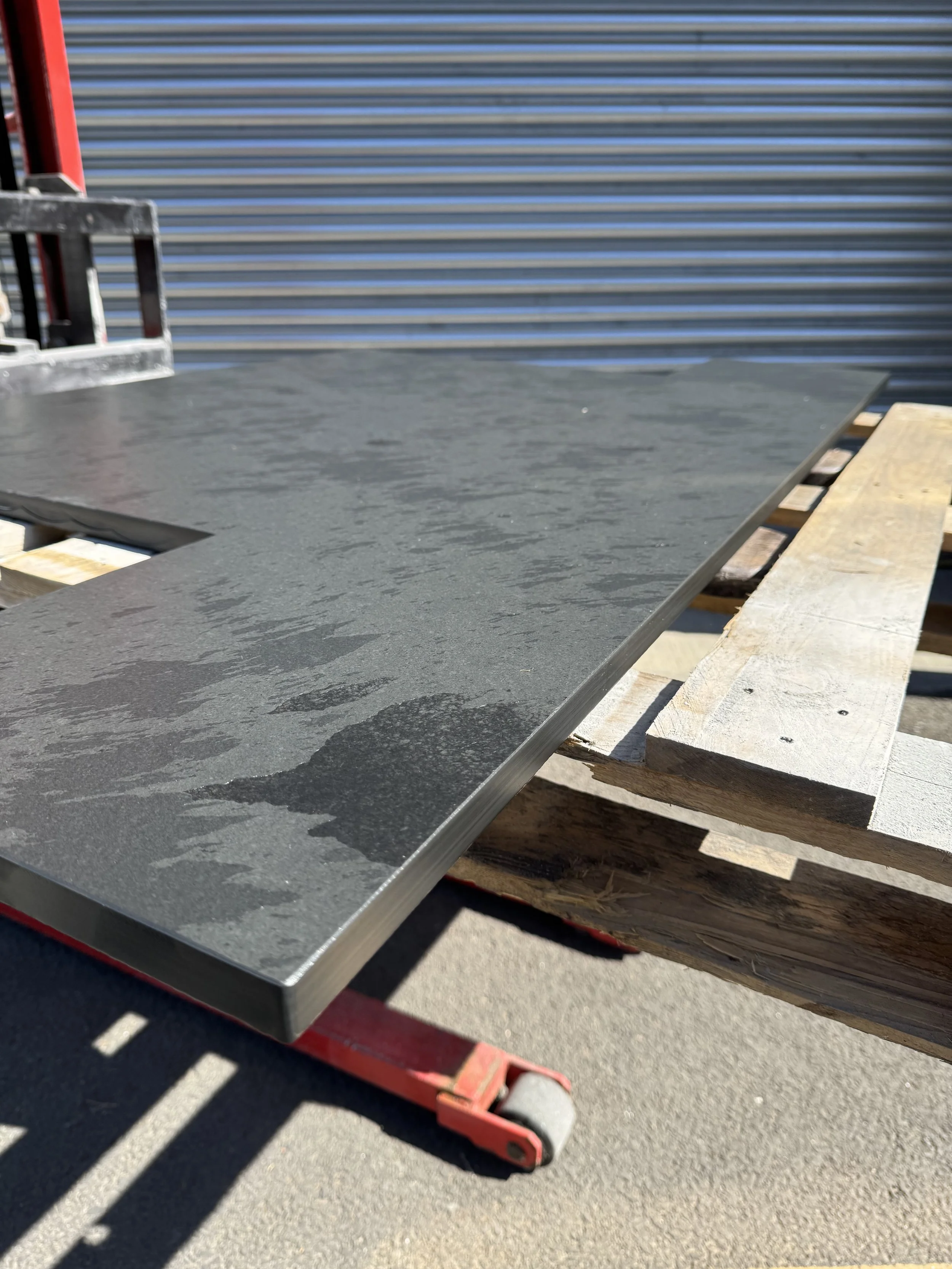 Close-up of a black textured stone slab resting on wooden pallets outdoors with metal siding in the background.