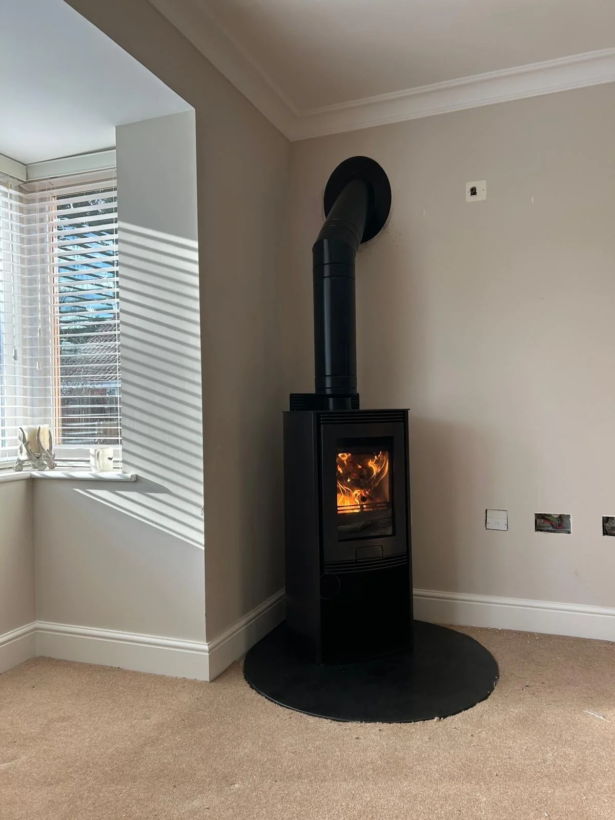 Black wood stove with glass door showing fire, connected to black pipe venting through the wall, in a living room with carpeted floor and light-colored walls.