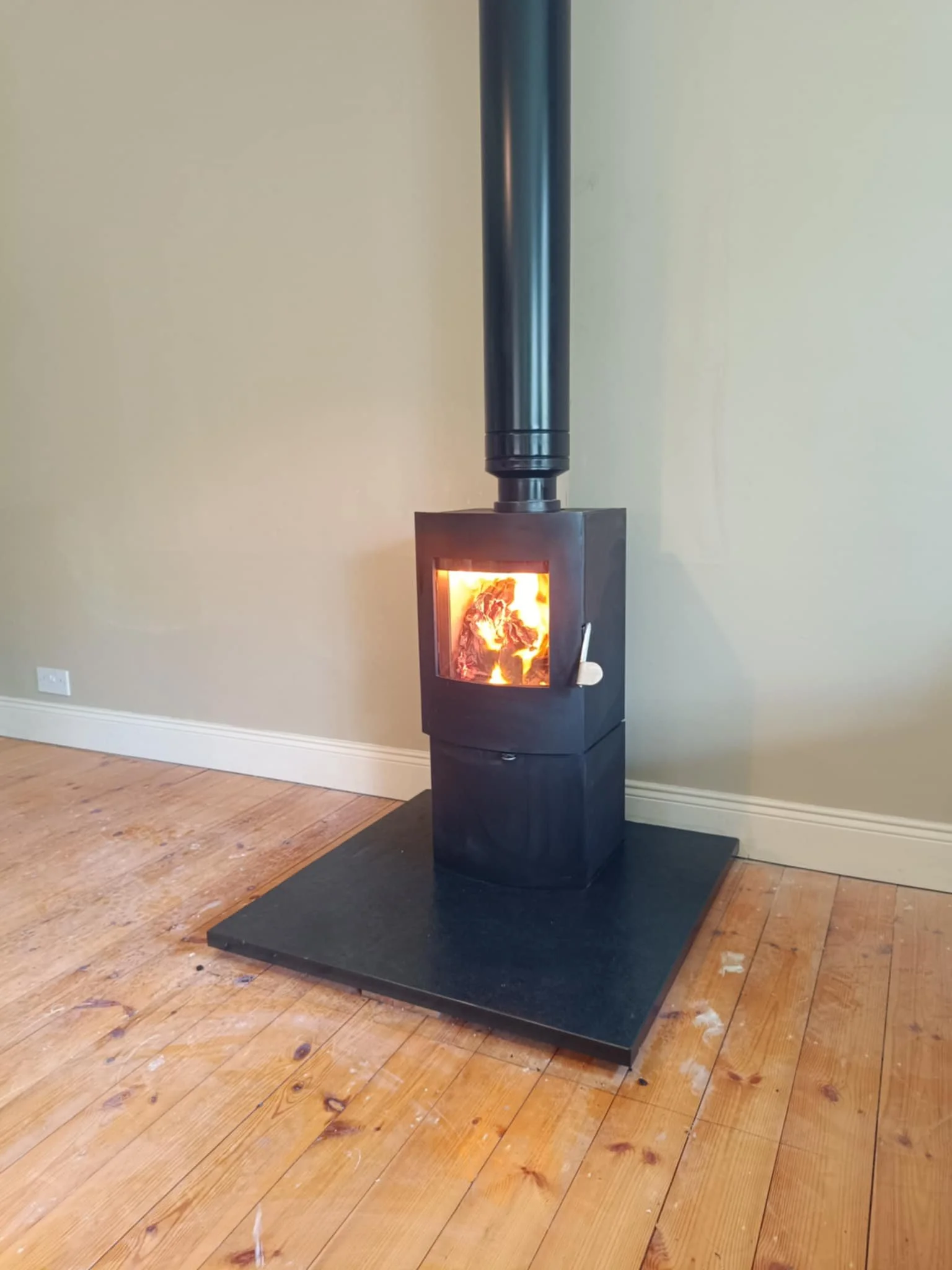 Black wood stove with a glass door showing fire inside, installed on a black hearth pad against a light-colored wall and wooden floor.